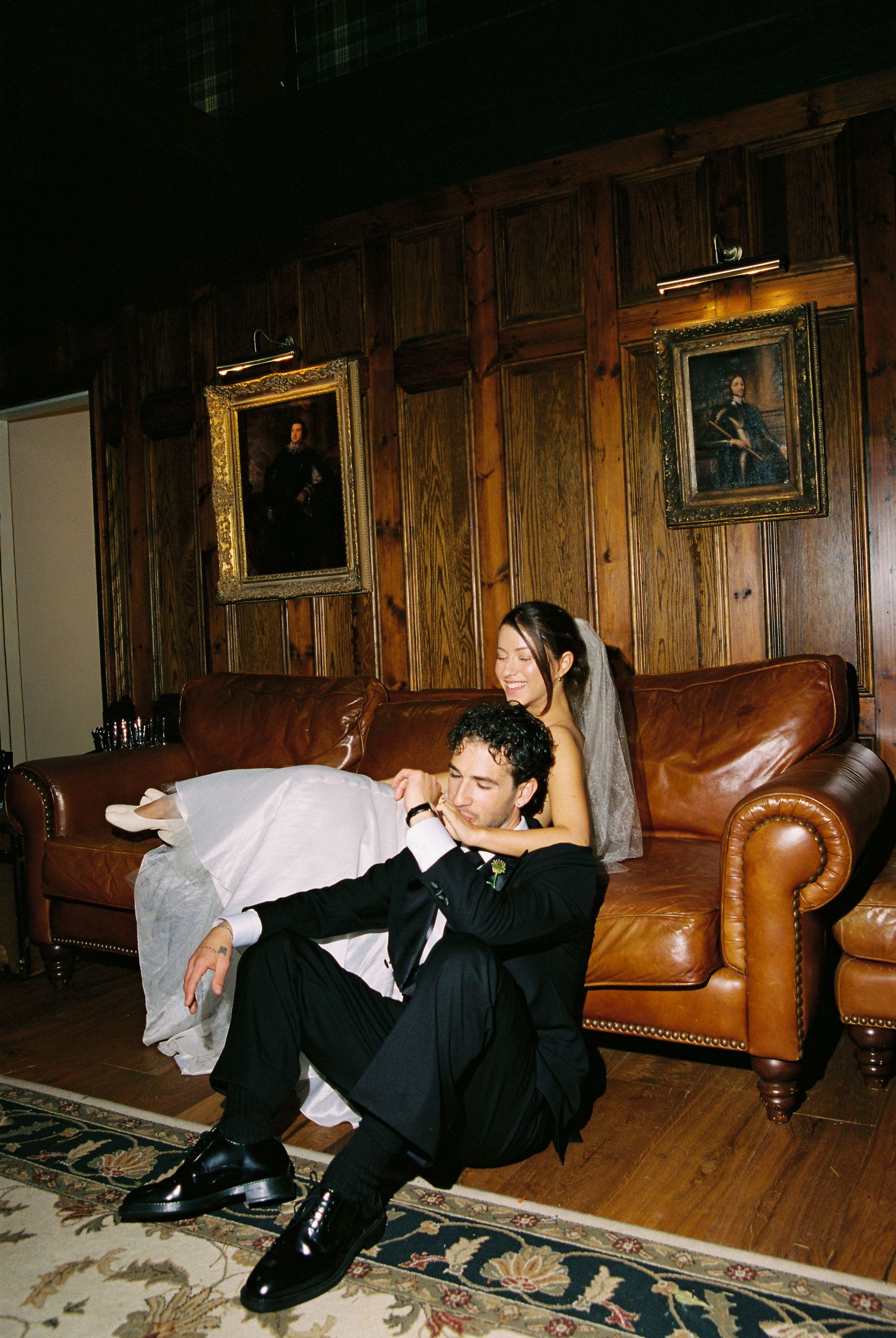 Bride and groom share an intimate moment on a leather sofa in a wood-paneled room with portrait paintings.