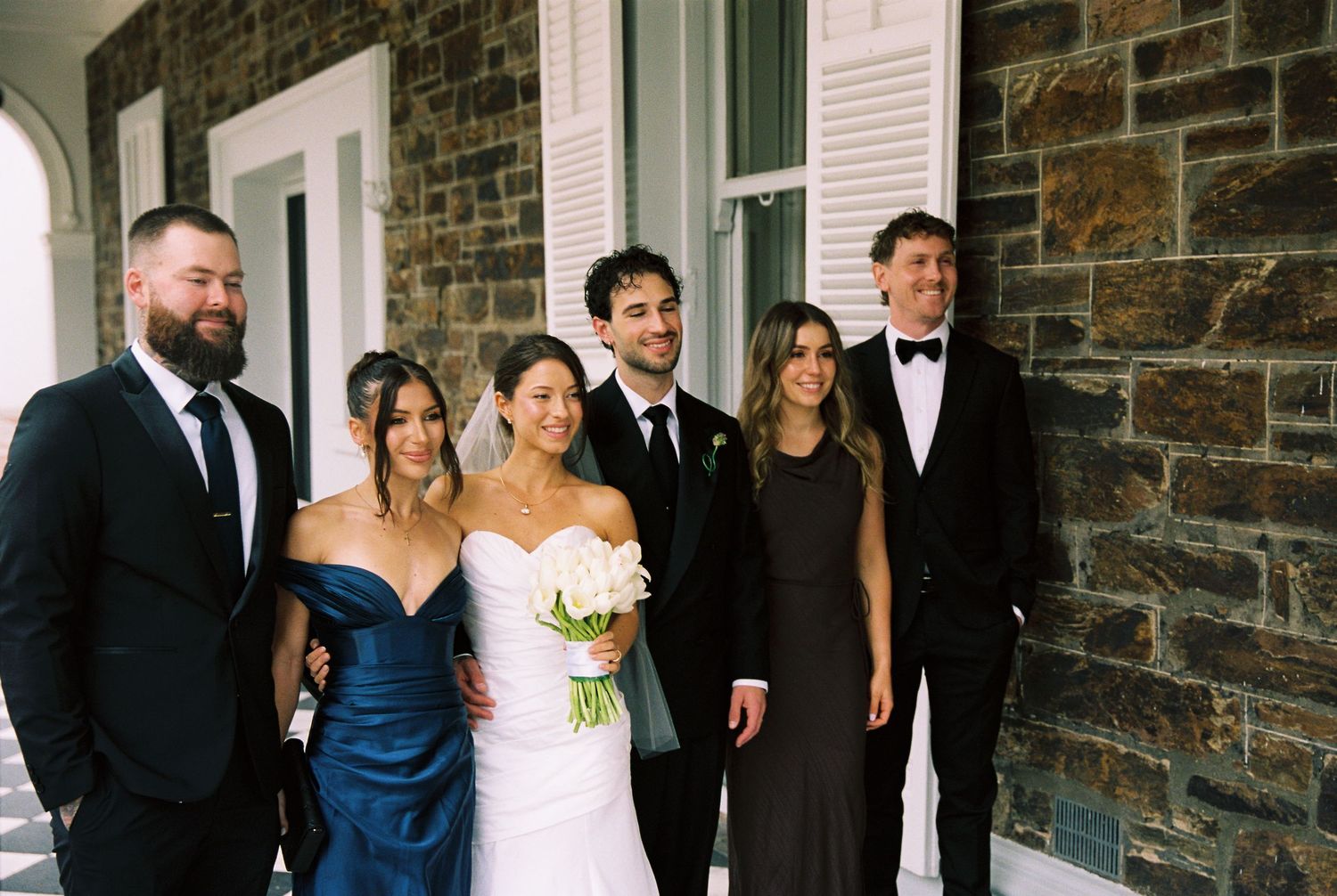 Wedding party posing together outside stone building, bride in white gown with bouquet, groom in black suit, guests in formal attire.