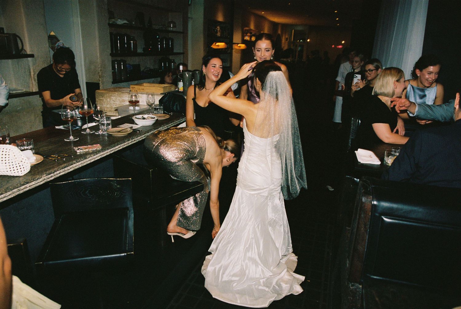 Bride in elegant white wedding gown with veil surrounded by guests celebrating at a wedding reception.
