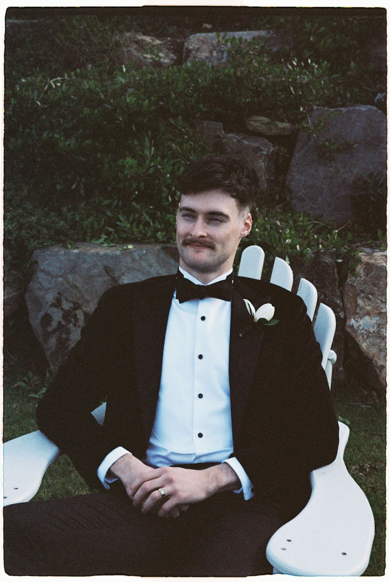 Groom in black tuxedo with bow tie sitting relaxed on white chair outdoors.