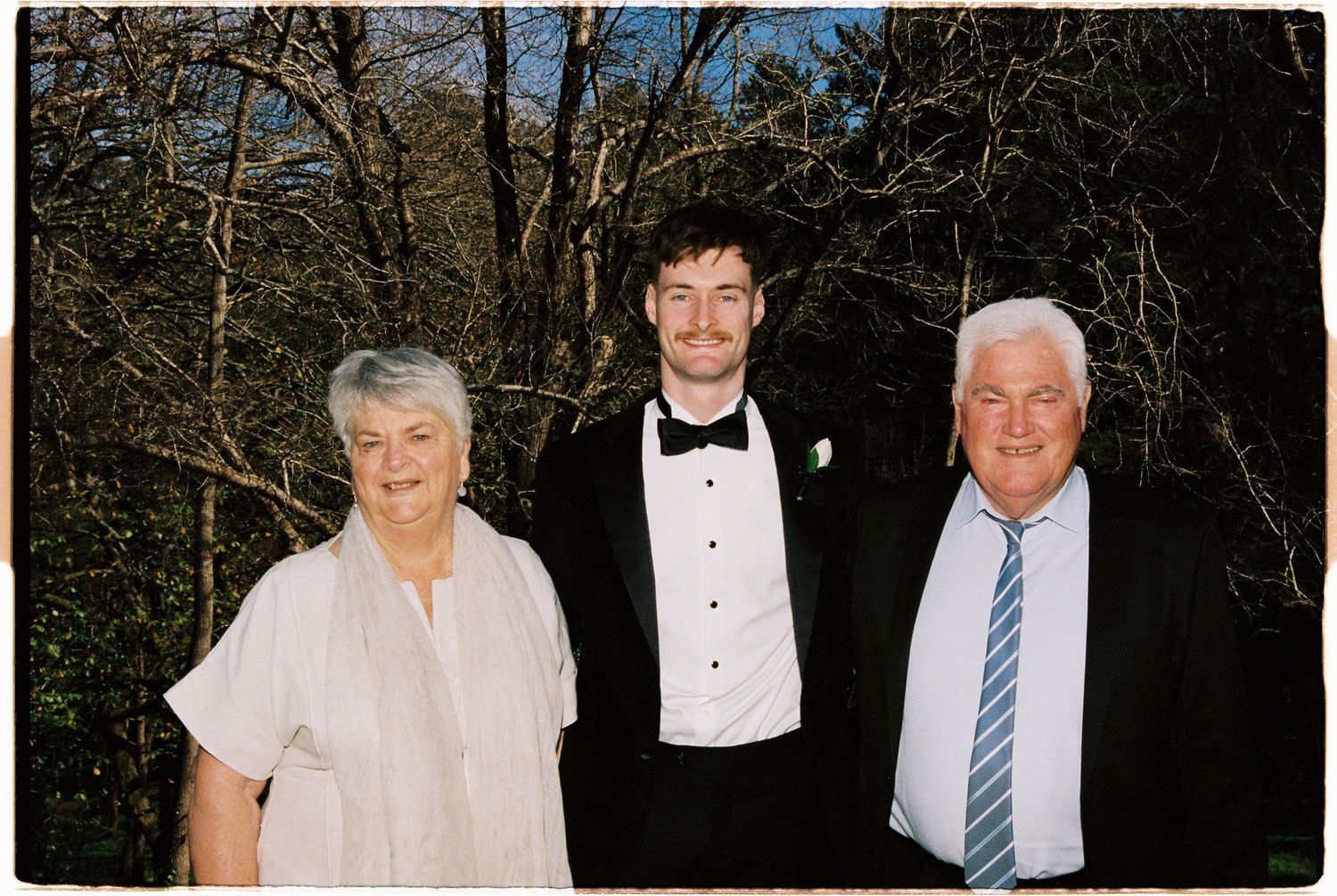 Family portrait of three people outdoors, young man in tuxedo with bow tie flanked by older woman and man in formal attire.