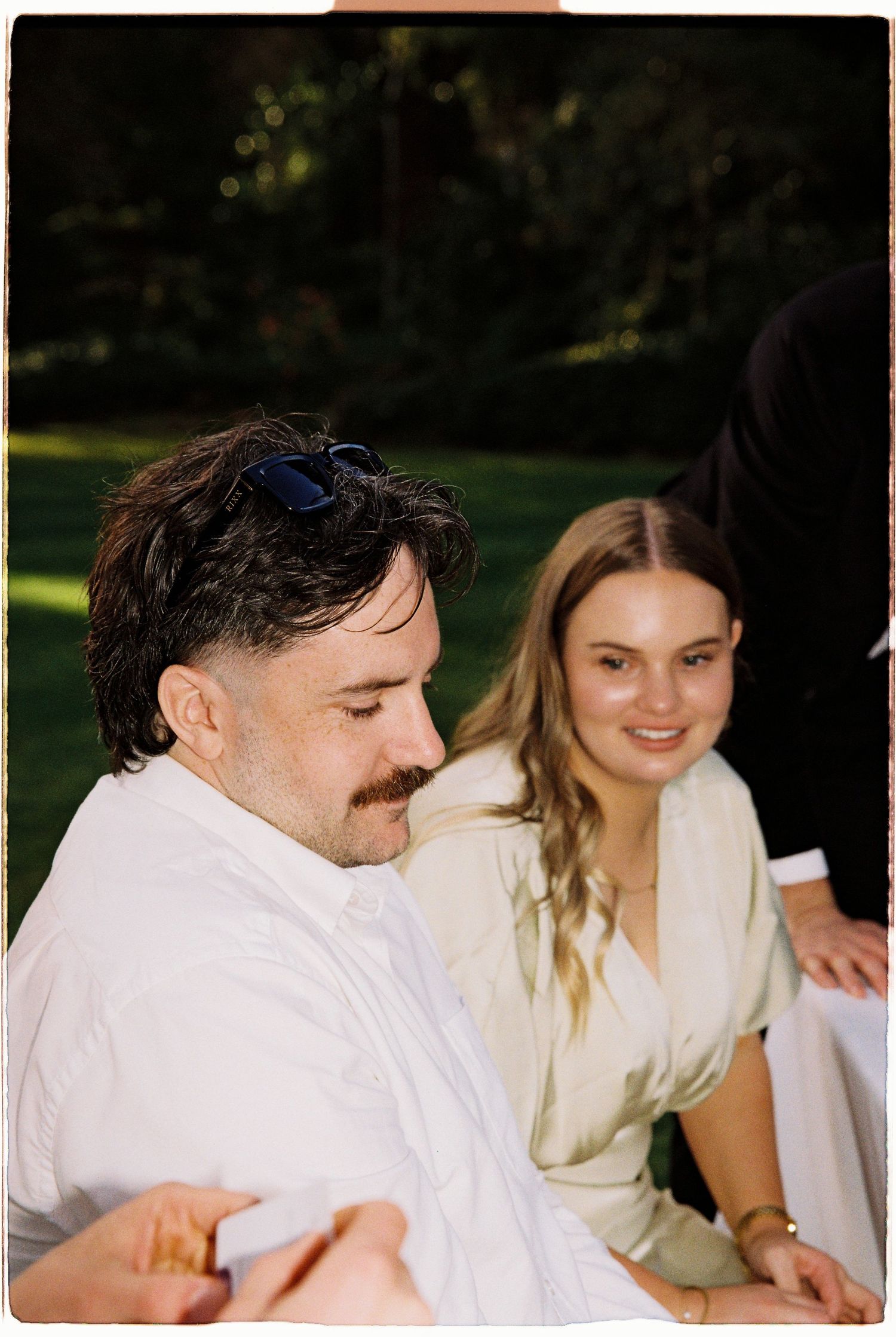 A man with sunglasses on his head and a mustache sits outdoors with a smiling blonde woman at a wedding reception.