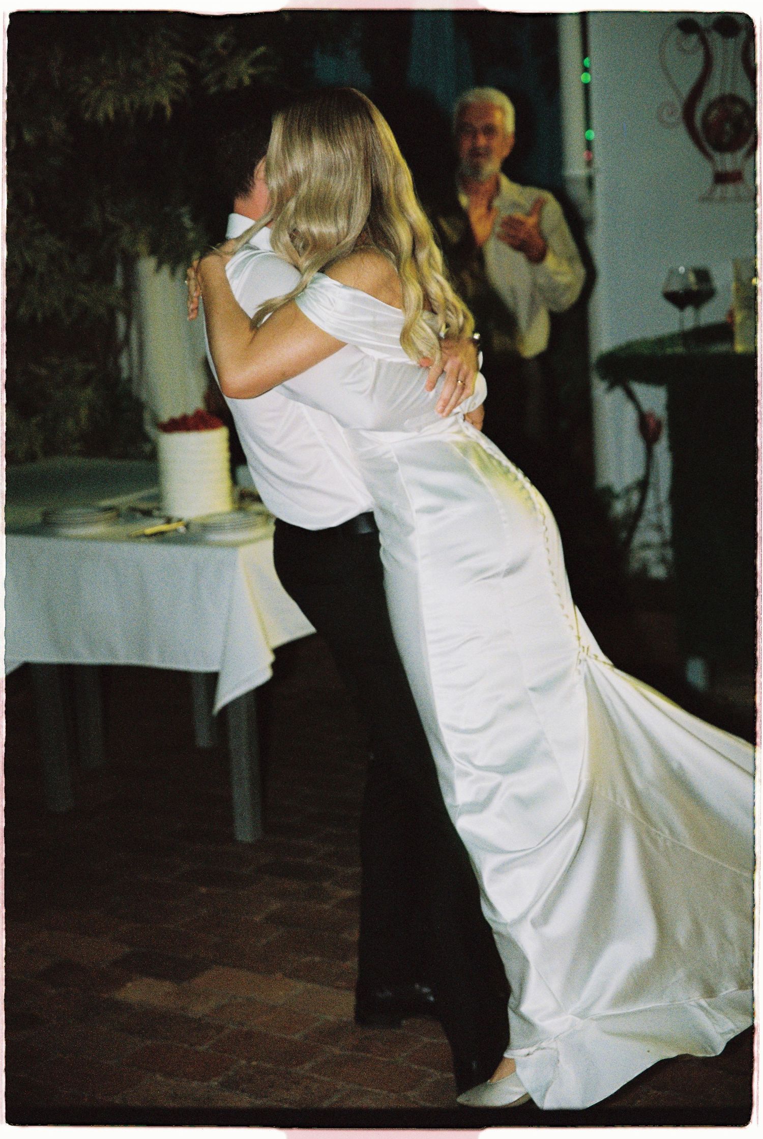 Bride and groom embracing during first dance at wedding reception with guests watching in background.