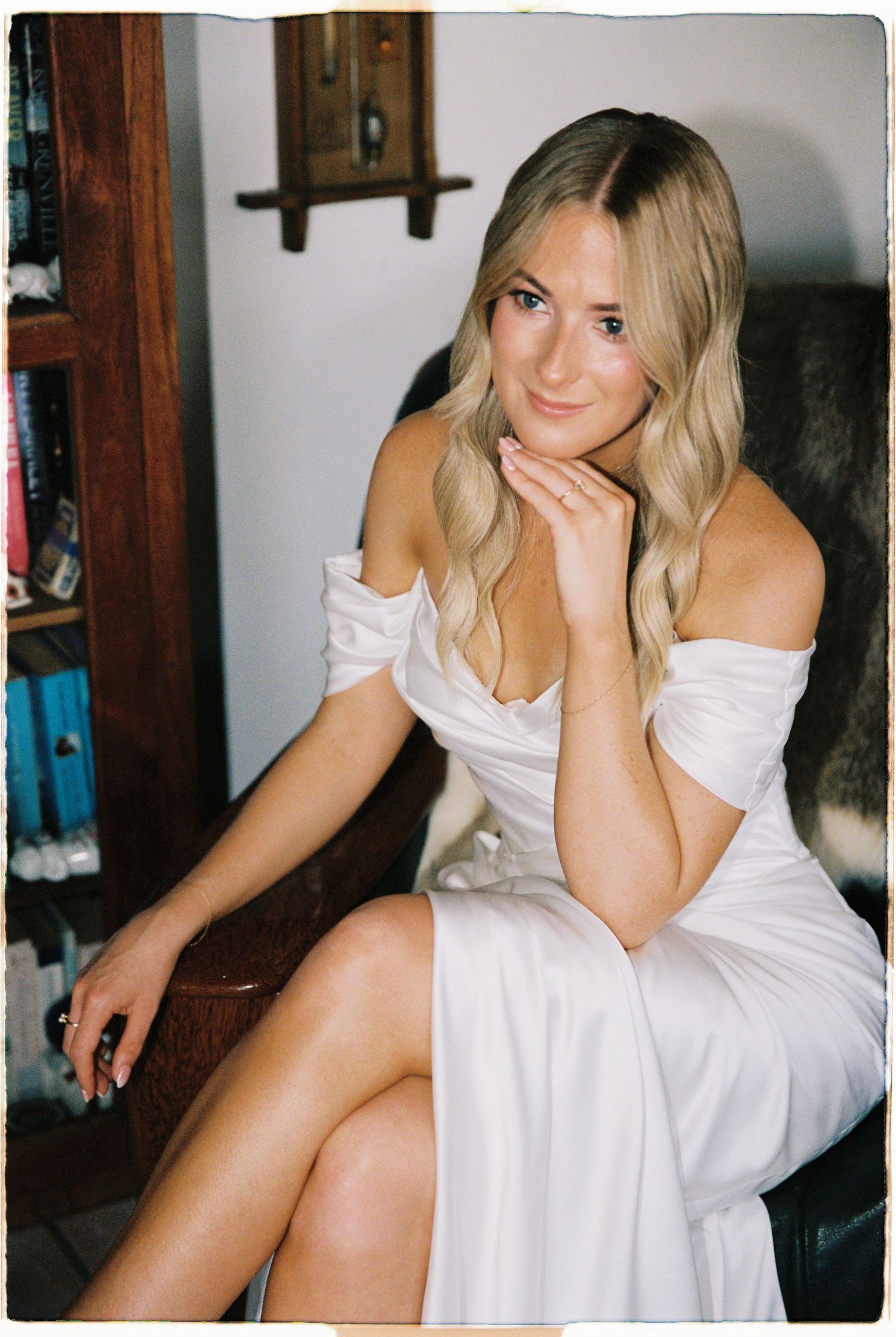 Blonde bride in off-shoulder white wedding dress seated in chair posing elegantly before ceremony.