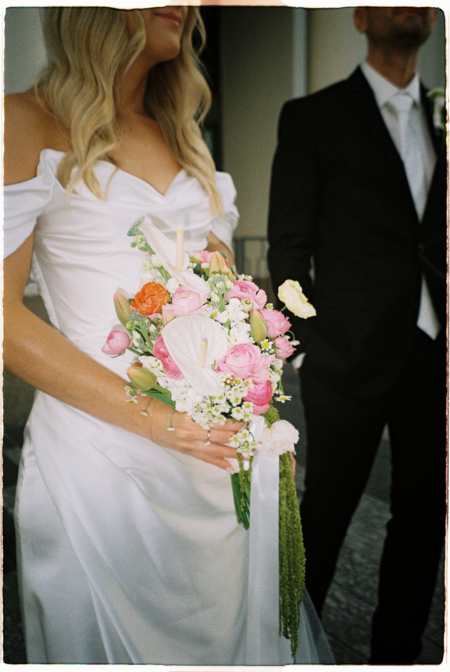 Bride in off-shoulder white gown holds a vibrant pink and white floral bouquet beside suited groom.