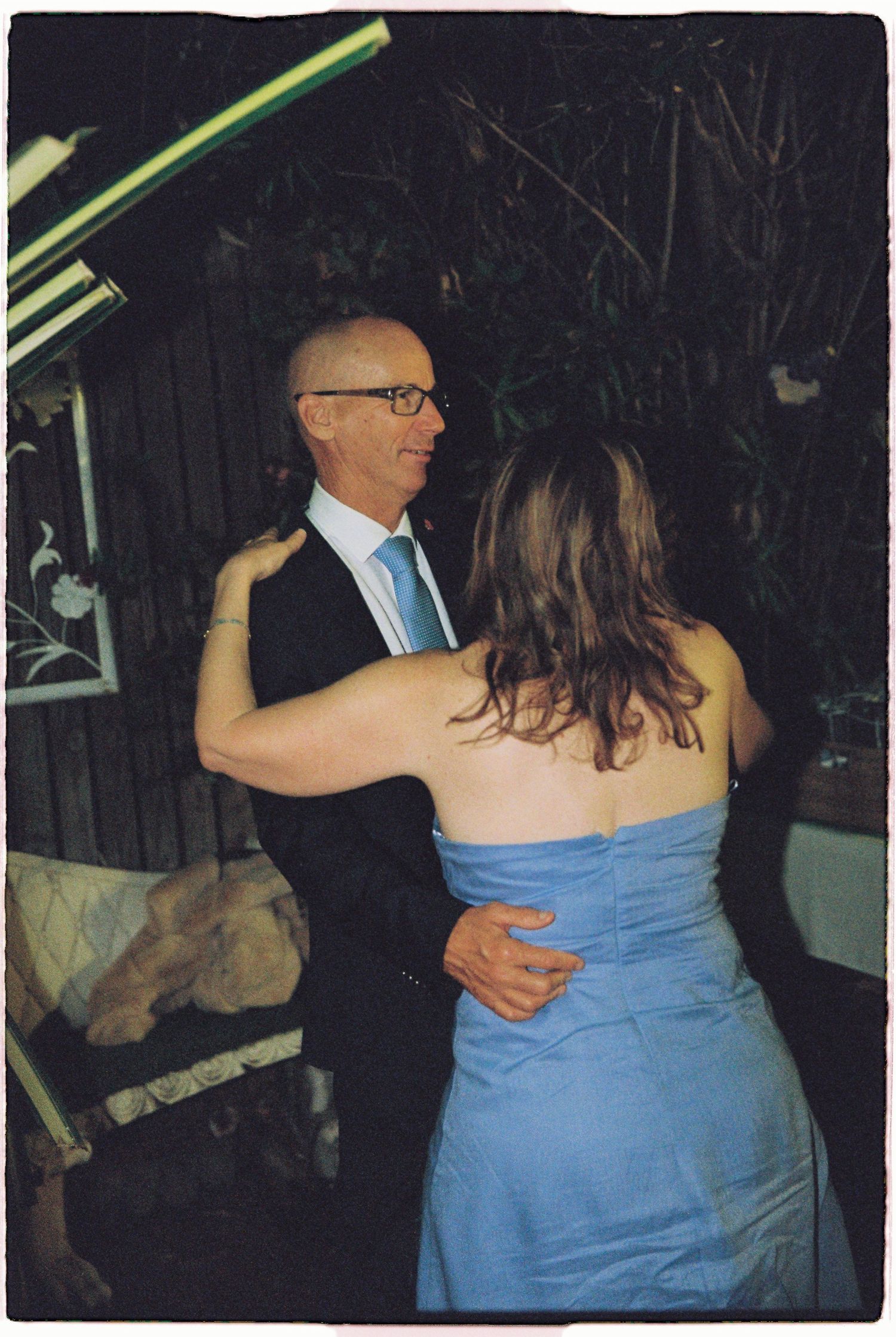 A bald man in glasses and suit dances with a woman in a blue strapless dress at an outdoor wedding reception.