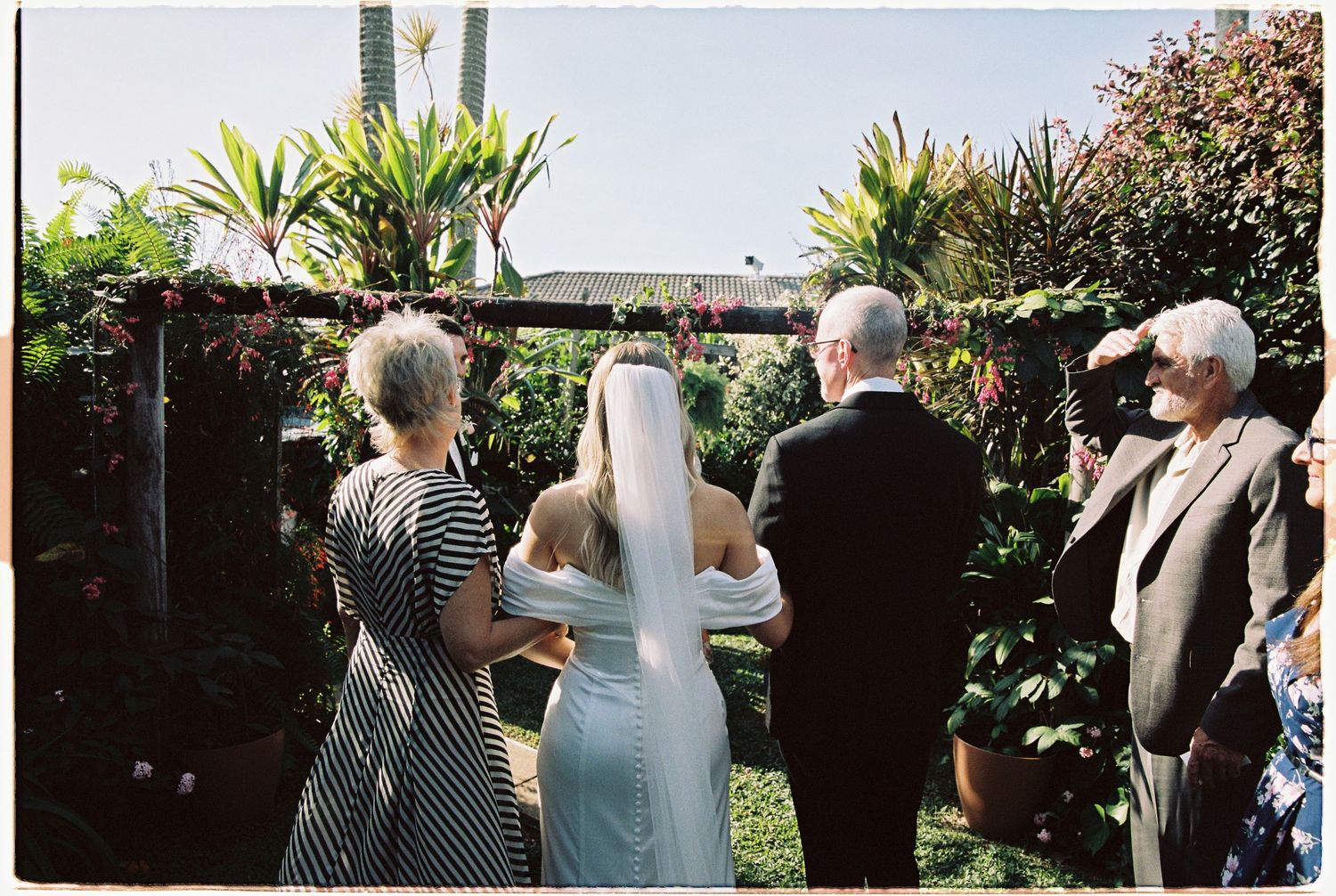 Bride in white gown and veil walks with family toward a lush tropical garden wedding ceremony arch.