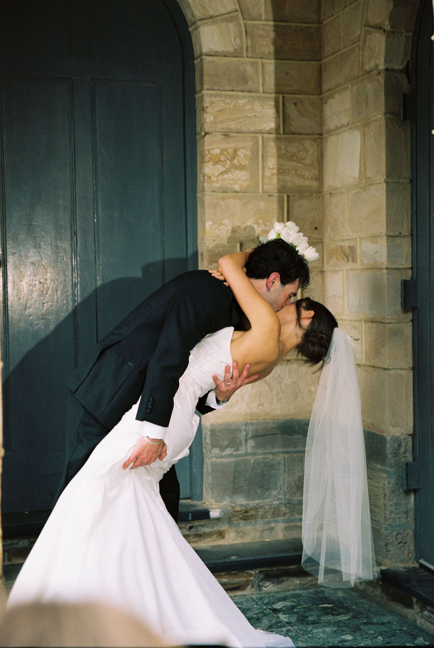Bride and groom sharing a romantic dip kiss in front of a stone church doorway on their wedding day.