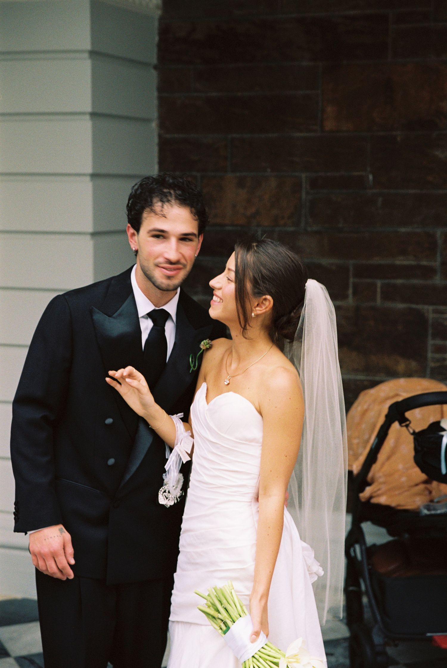 Bride and groom posing together outside church, bride in strapless white gown holding tulip bouquet.