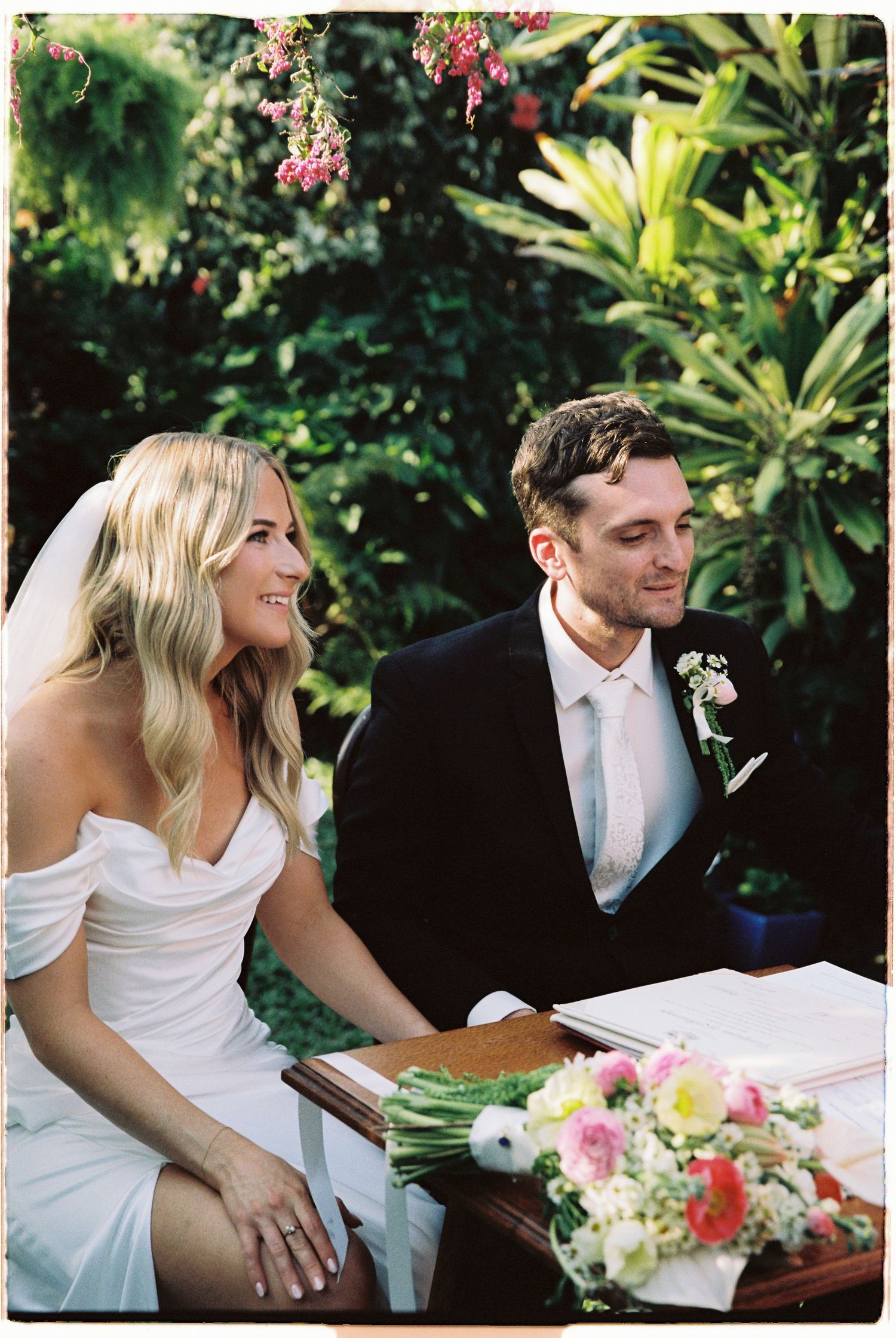 Bride and groom sit together at outdoor wedding ceremony signing table with floral bouquet.