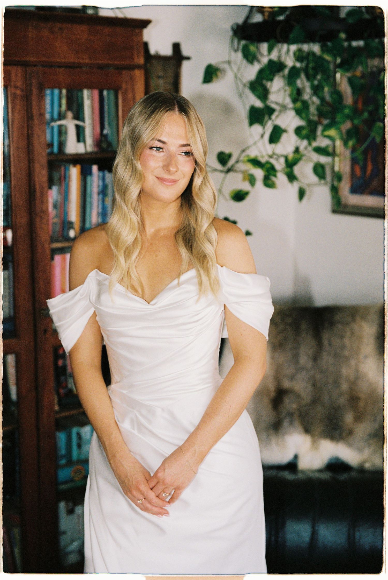 Blonde bride in elegant off-shoulder white wedding gown posing indoors near a bookshelf.