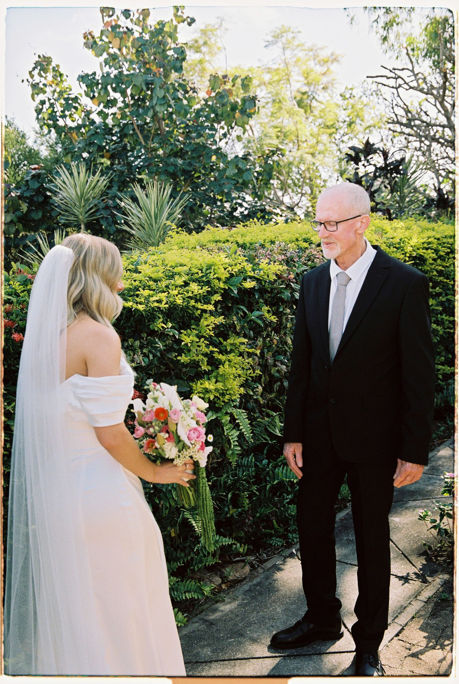 Bride in white veil and off-shoulder gown shares first look moment with father outdoors.