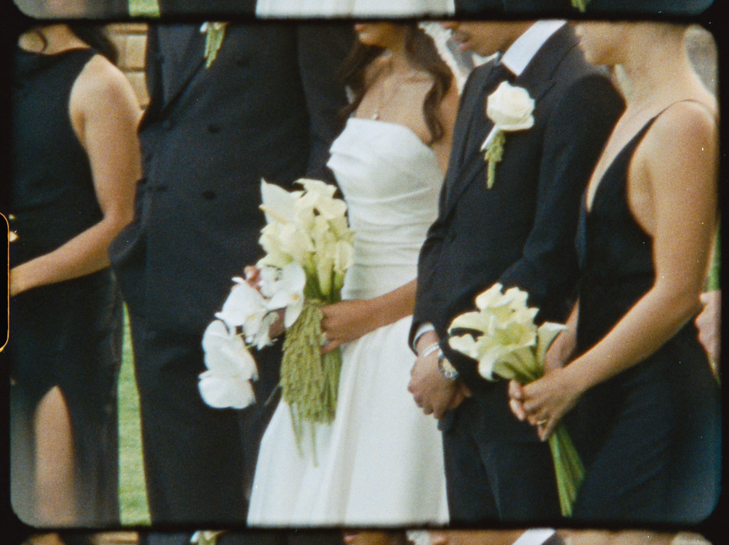 Wedding party in black attire surrounds bride in white gown holding white calla lily bouquet.