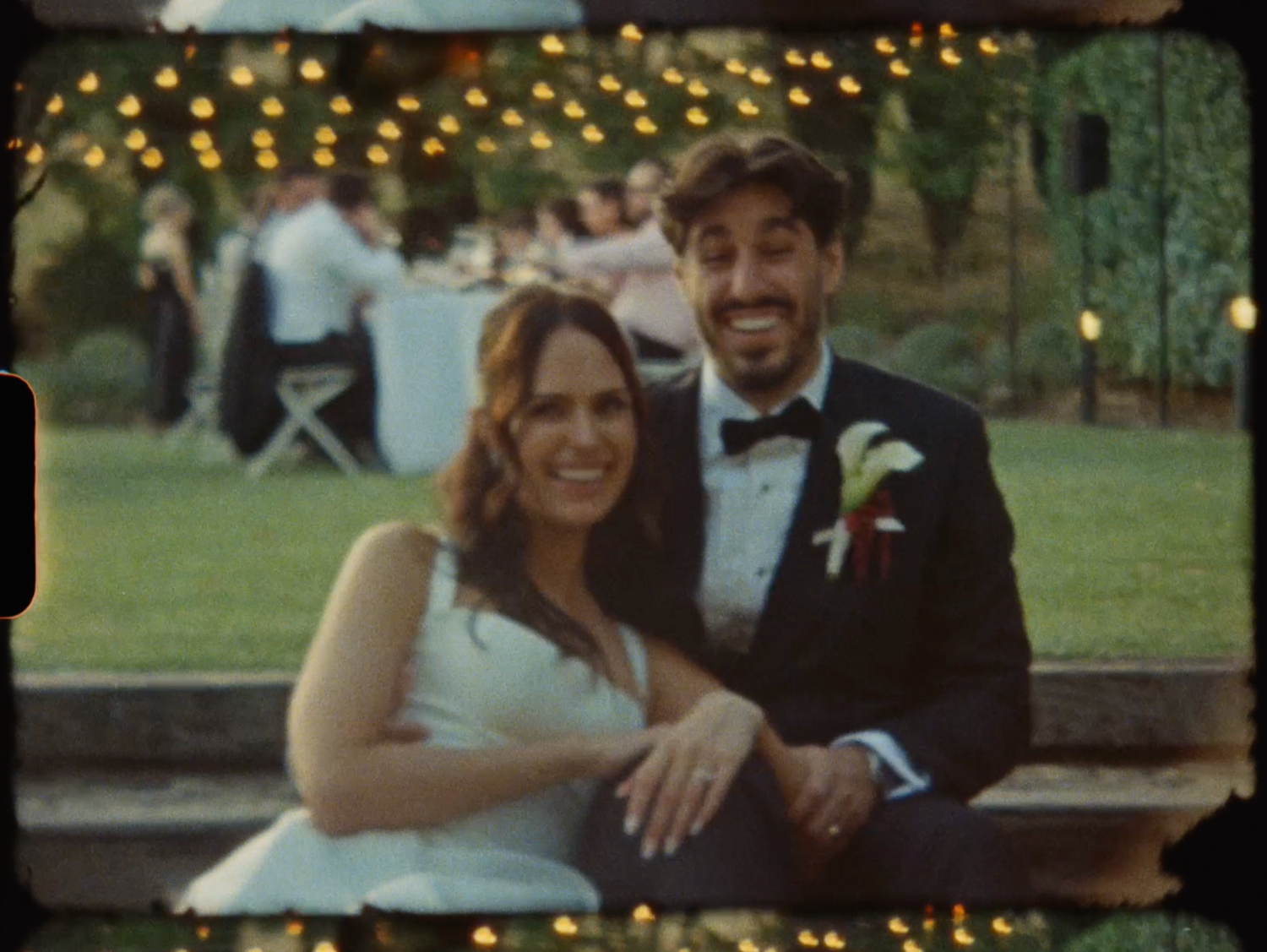 Smiling bride and groom pose together at outdoor evening wedding reception with warm string lights behind them.
