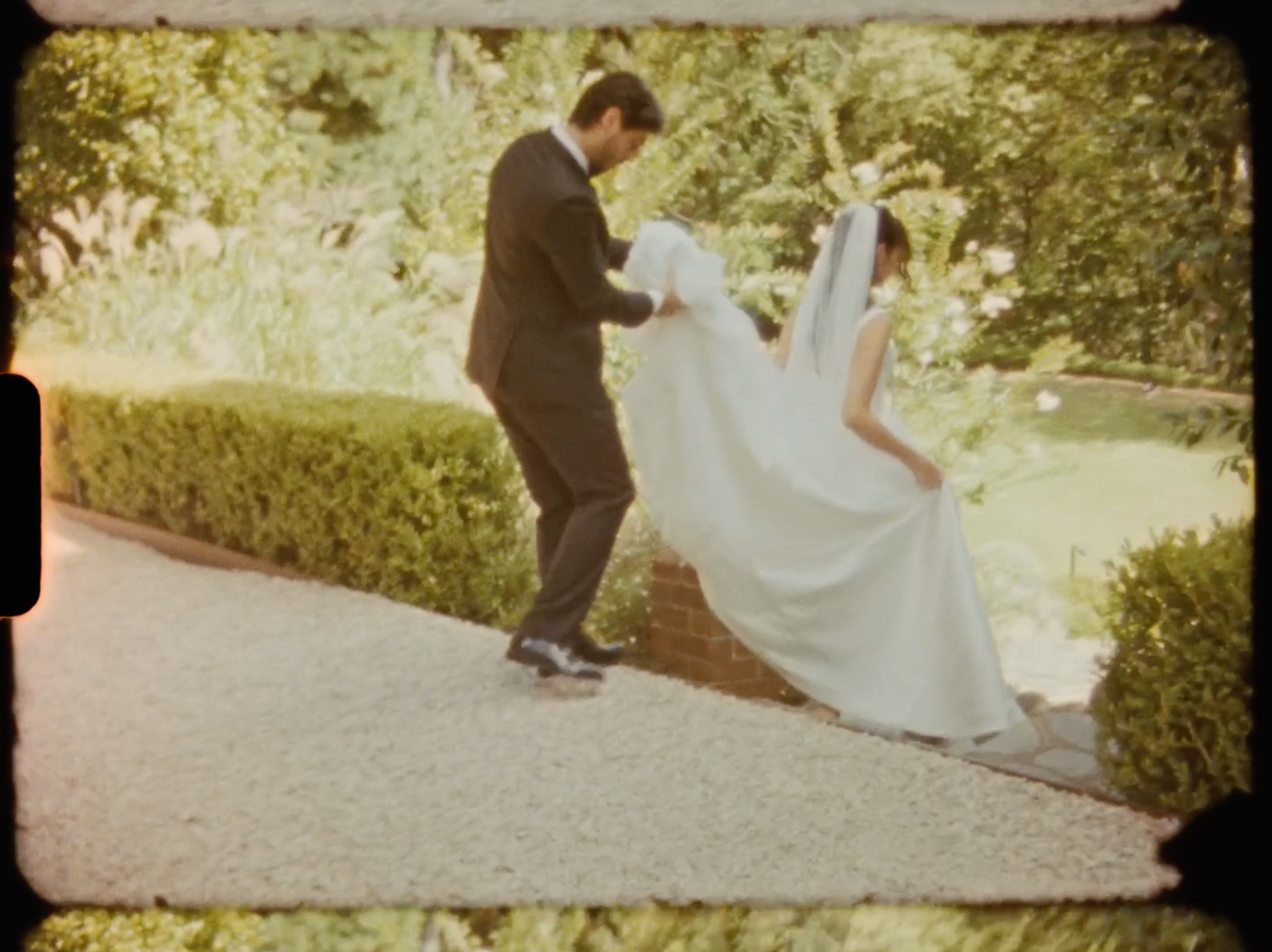 Groom in black tuxedo adjusting bride's white wedding gown and veil on garden pathway.