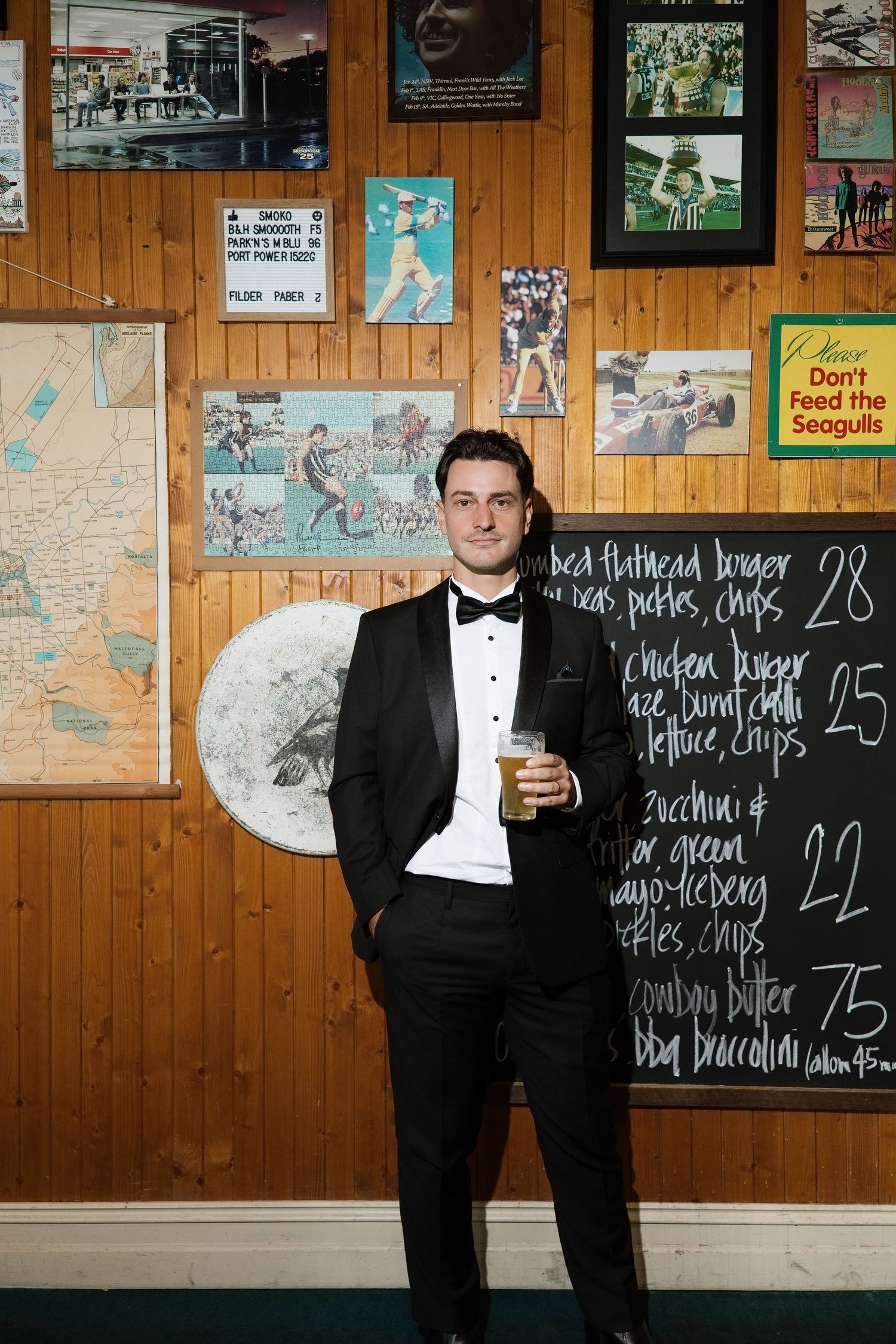 Groom in black tuxedo holding a drink, standing confidently in front of a wood-paneled pub wall.