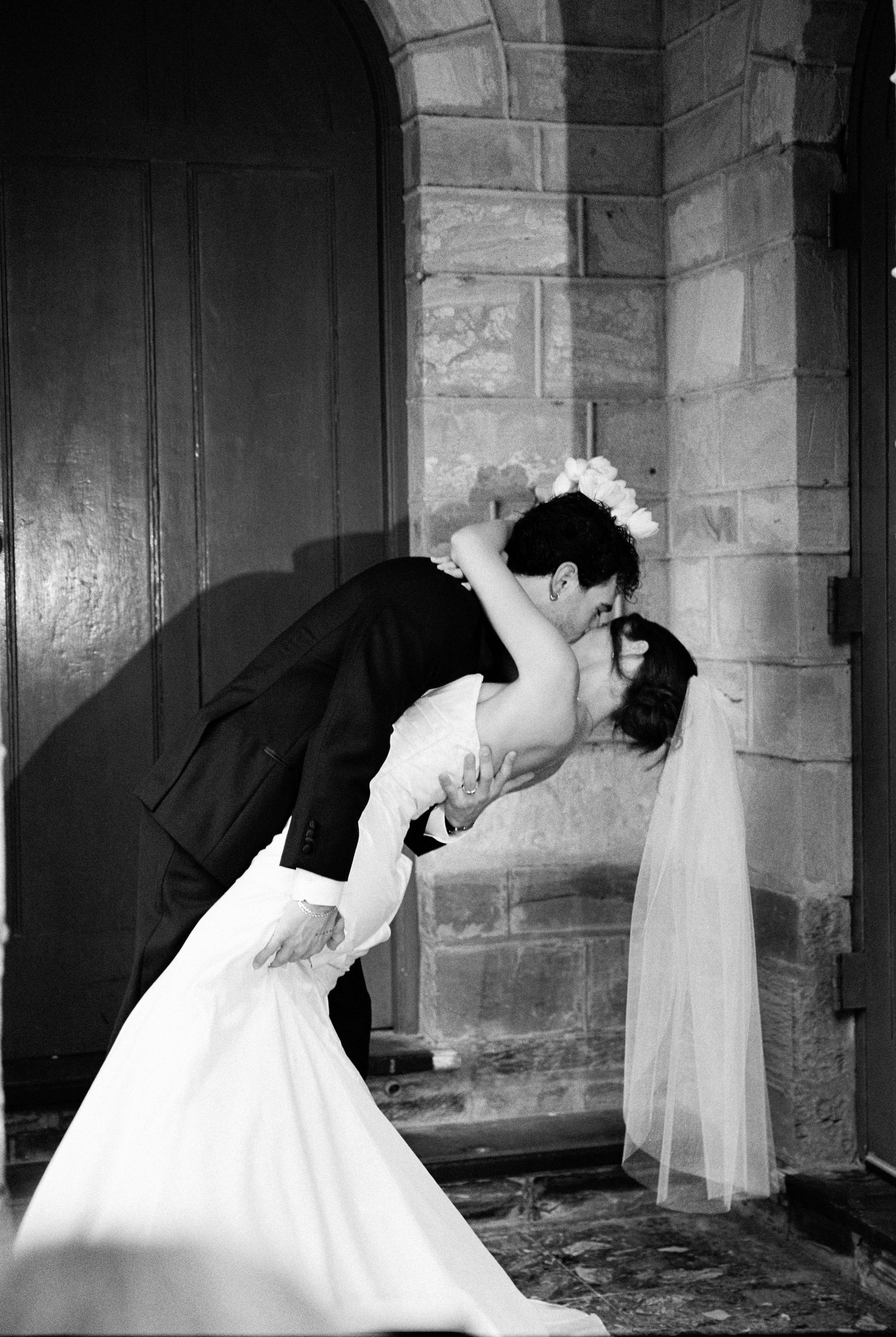 Bride and groom sharing a romantic dip kiss outside stone church doorway.