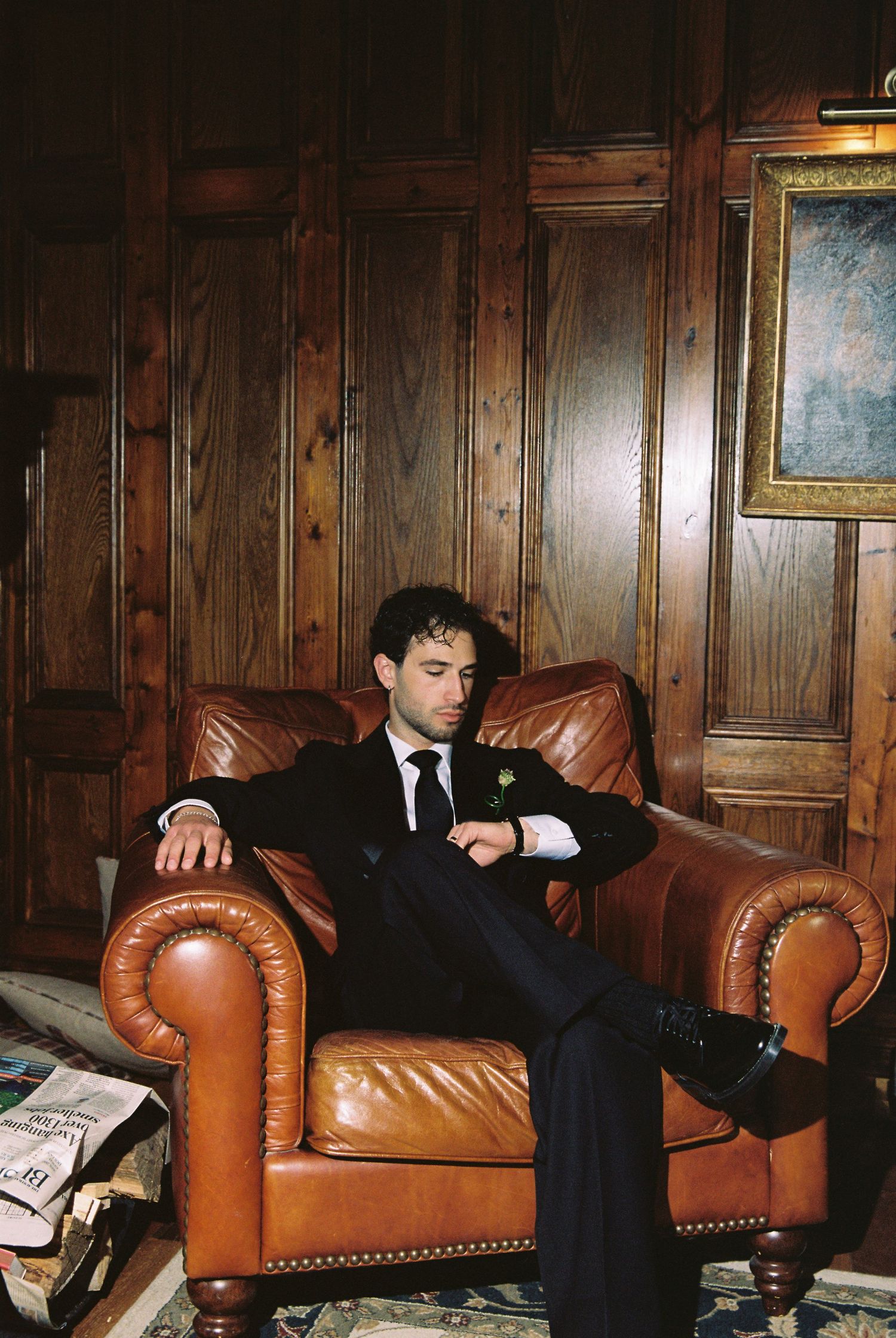 Groom in black tuxedo relaxing in brown leather armchair in a wood-paneled room checking his watch.