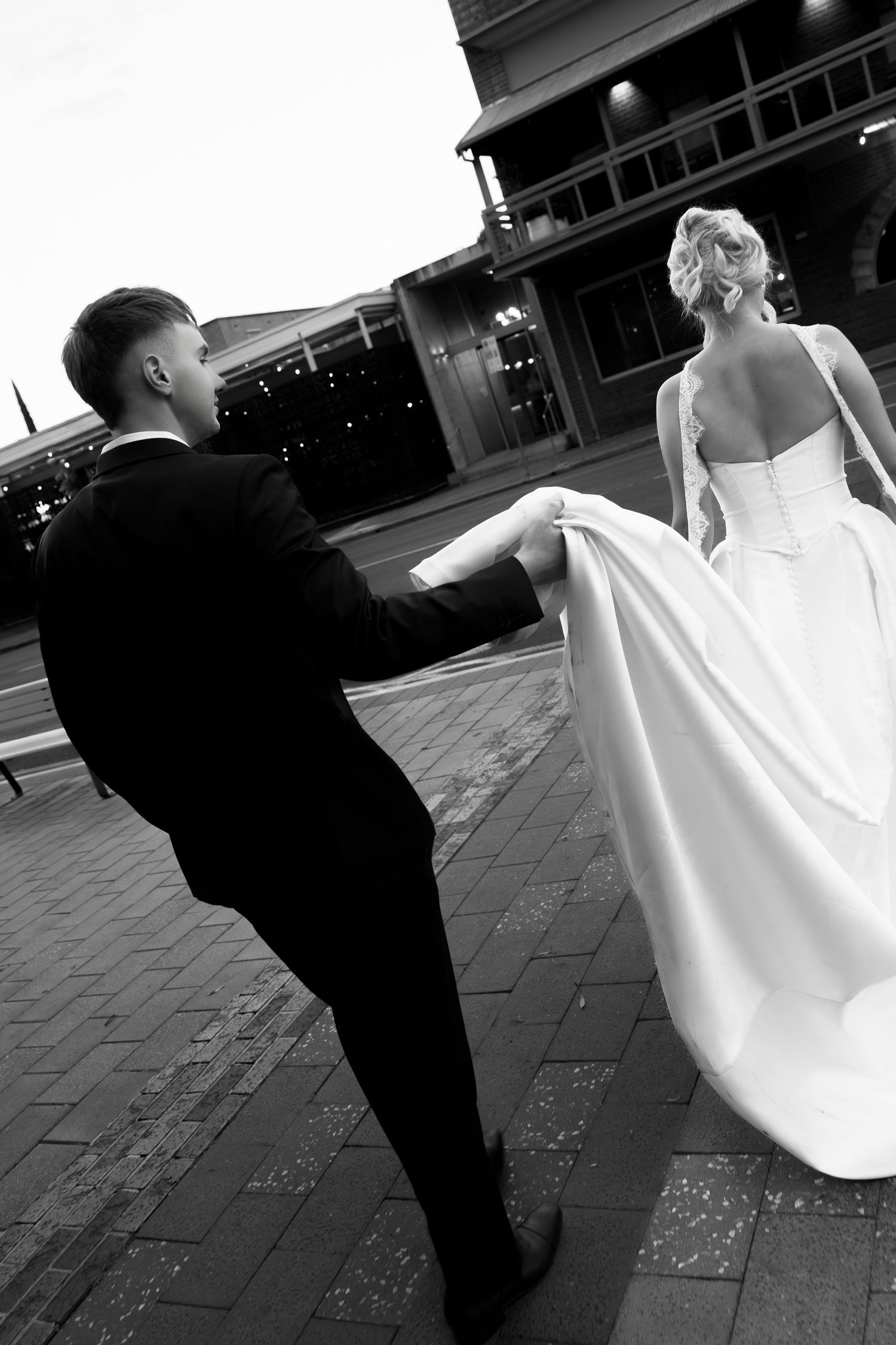 Black and white photo of groom holding bride's wedding dress train as they walk along a city street at dusk.