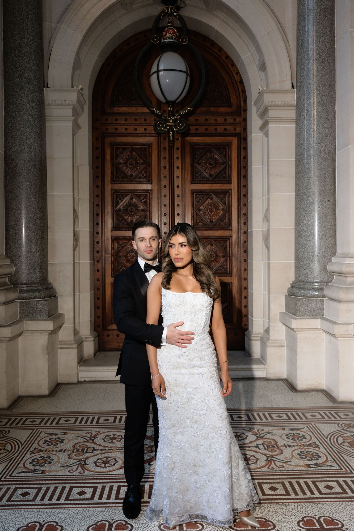 Elegant bride and groom posing in front of ornate wooden doors with marble columns on their wedding day.