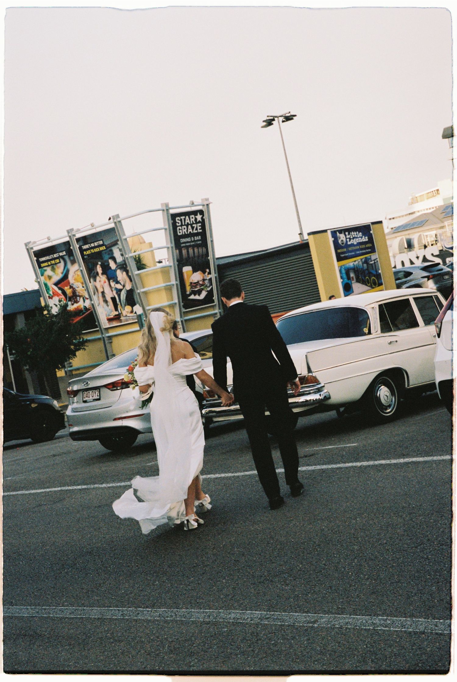 Bride and groom walking toward vintage white station wagon in parking lot.