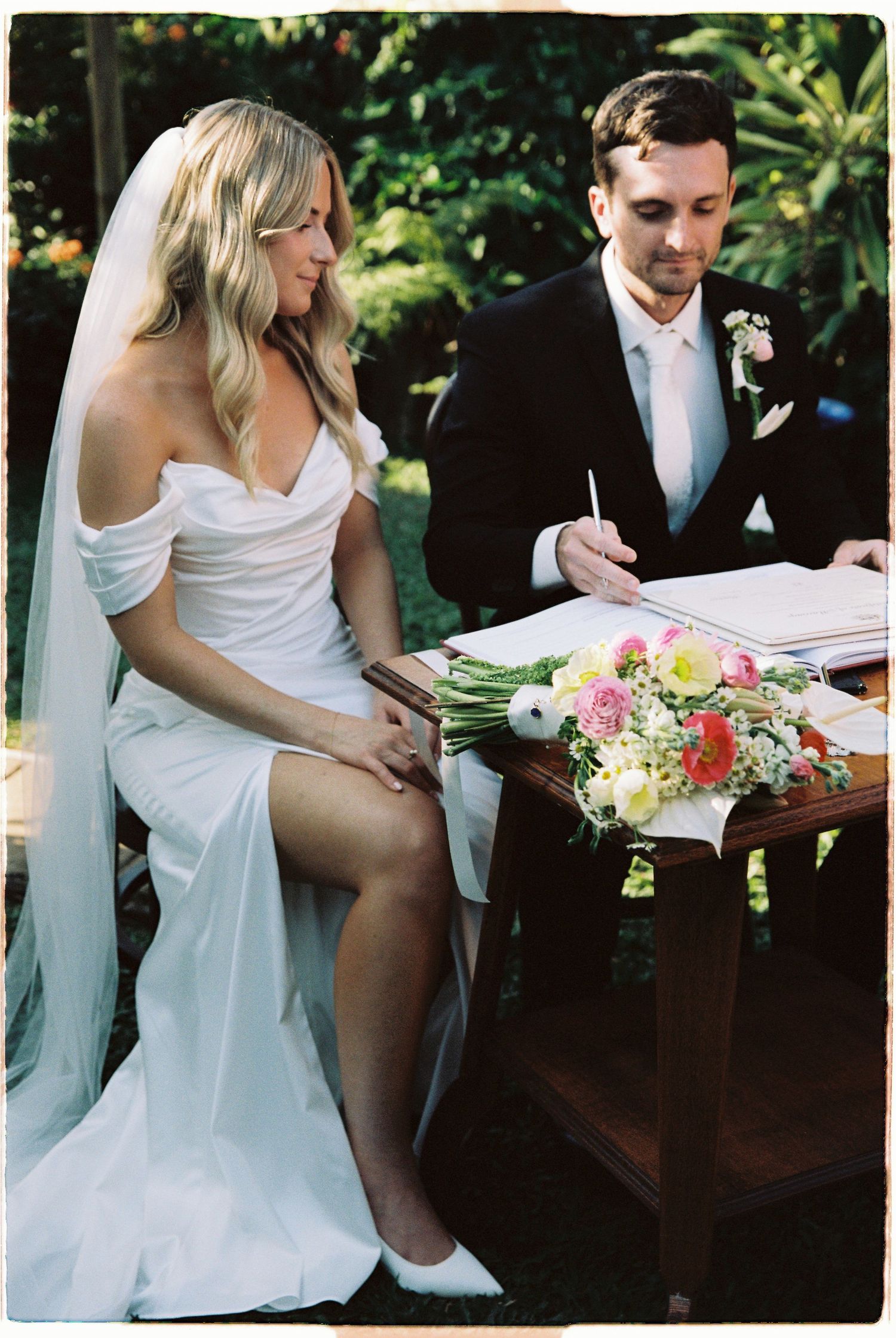 Bride and groom signing wedding documents at a wooden table adorned with colorful flowers during outdoor ceremony.