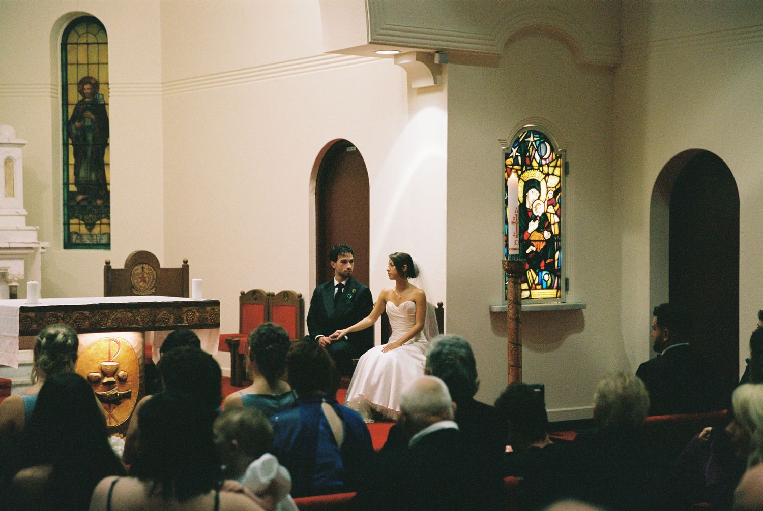 Bride and groom stand before guests in small church with stained glass windows during wedding ceremony.