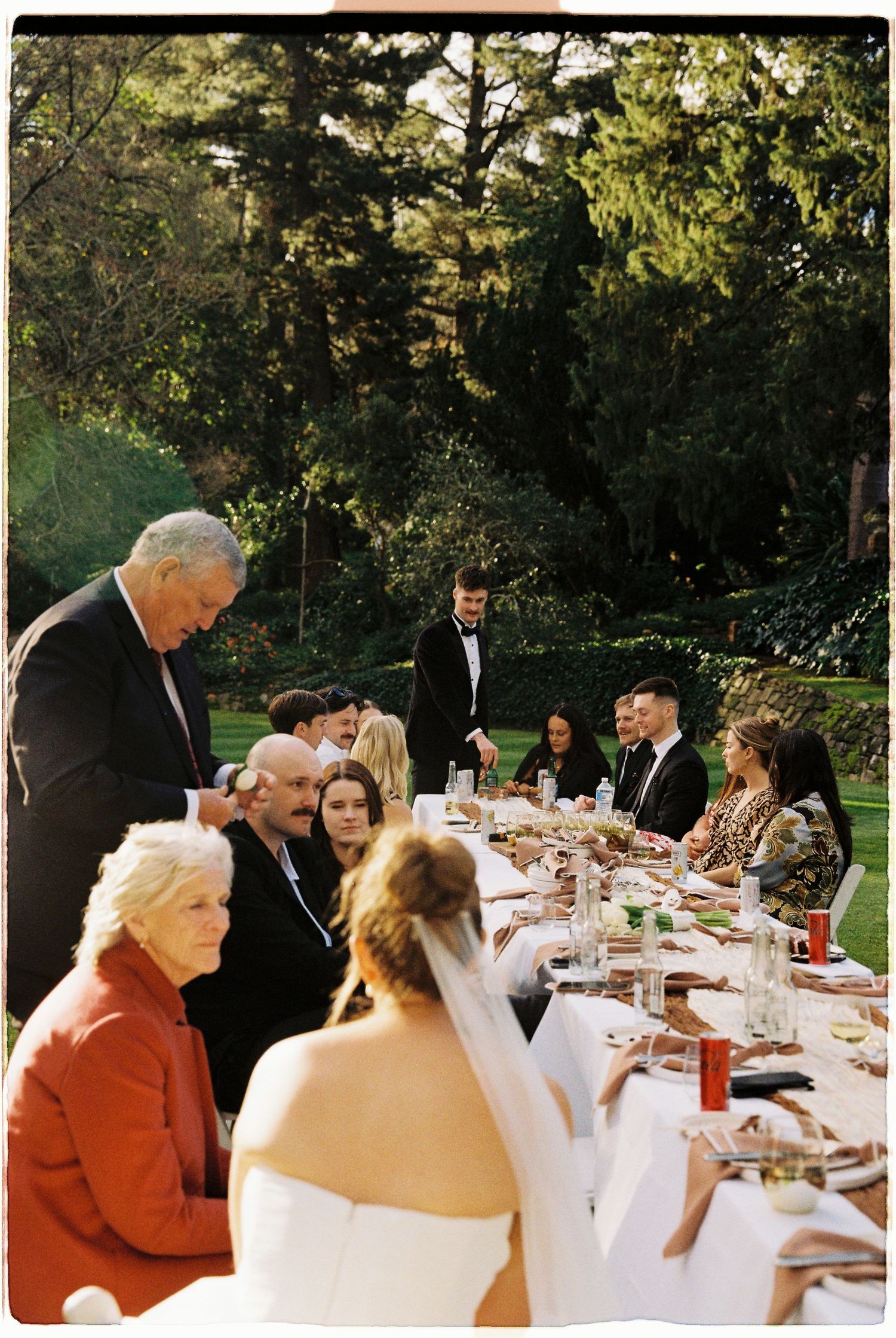 Wedding guests seated at long outdoor reception table while man gives speech in garden setting.