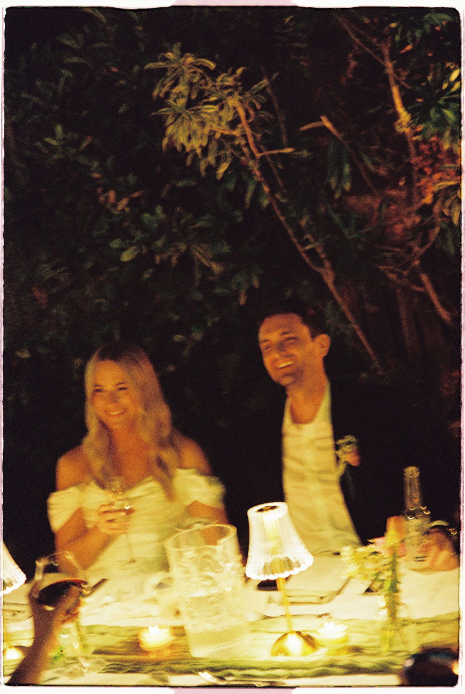 Bride and groom laughing together at candlelit outdoor wedding reception dinner table at night.