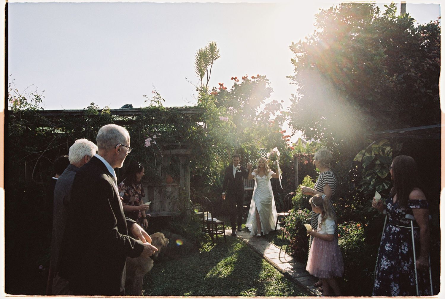 Outdoor garden wedding ceremony with guests gathered in golden sunlight amid lush greenery.