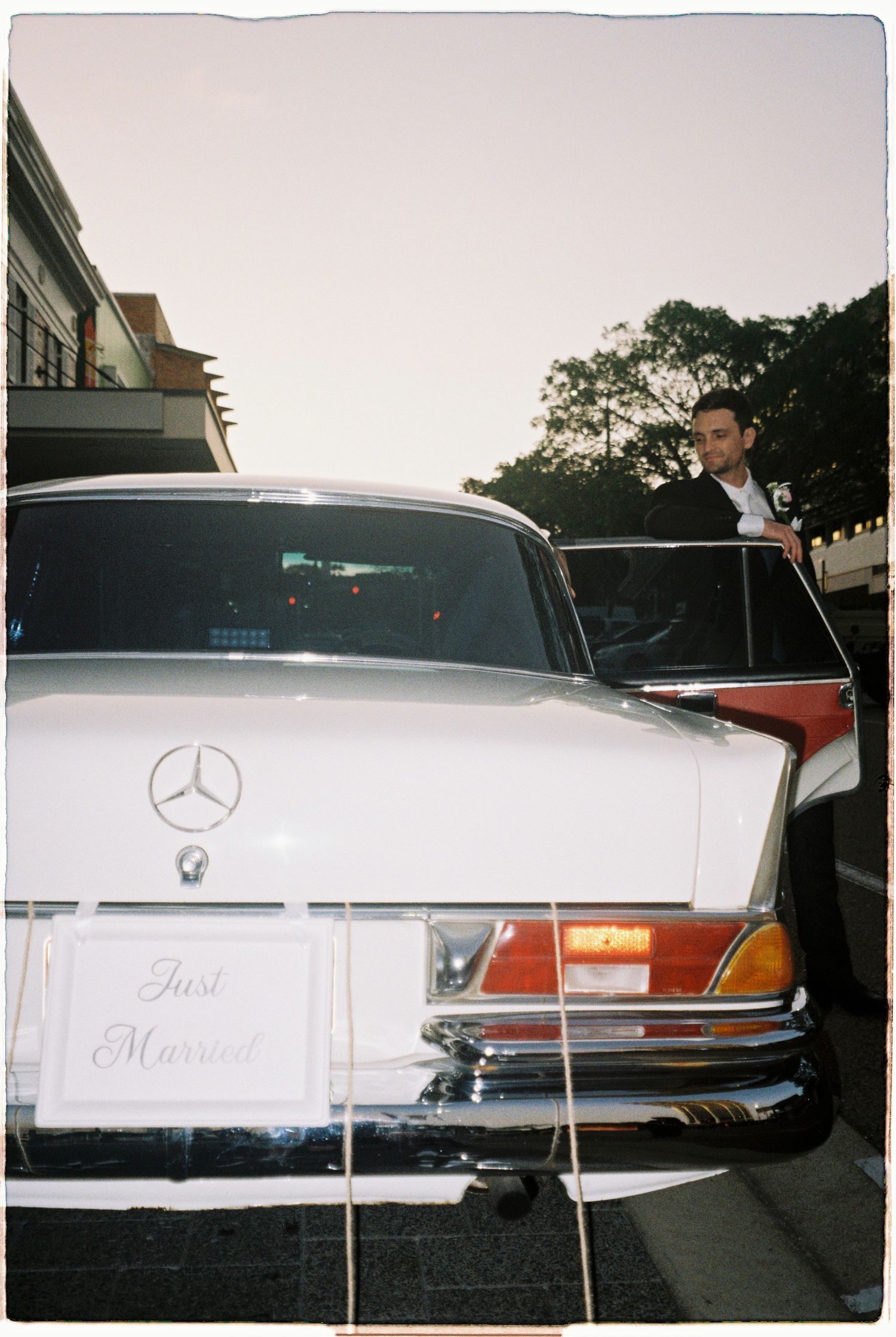 A vintage white Mercedes-Benz wedding car with a Just Married sign, groom standing behind it.