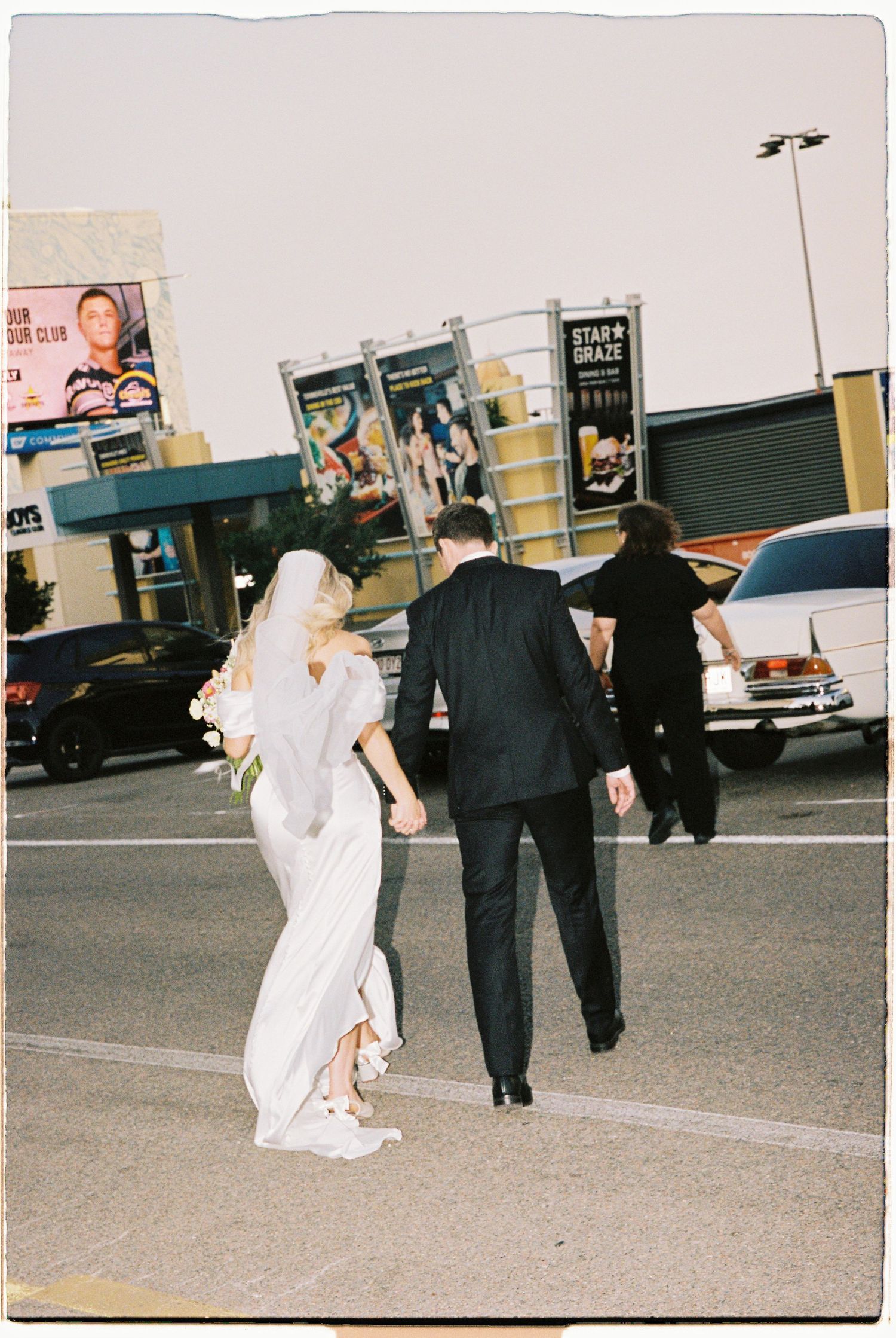 Bride and groom cross a parking lot hand in hand, bride in white gown with veil, groom in black suit, vintage car behind.