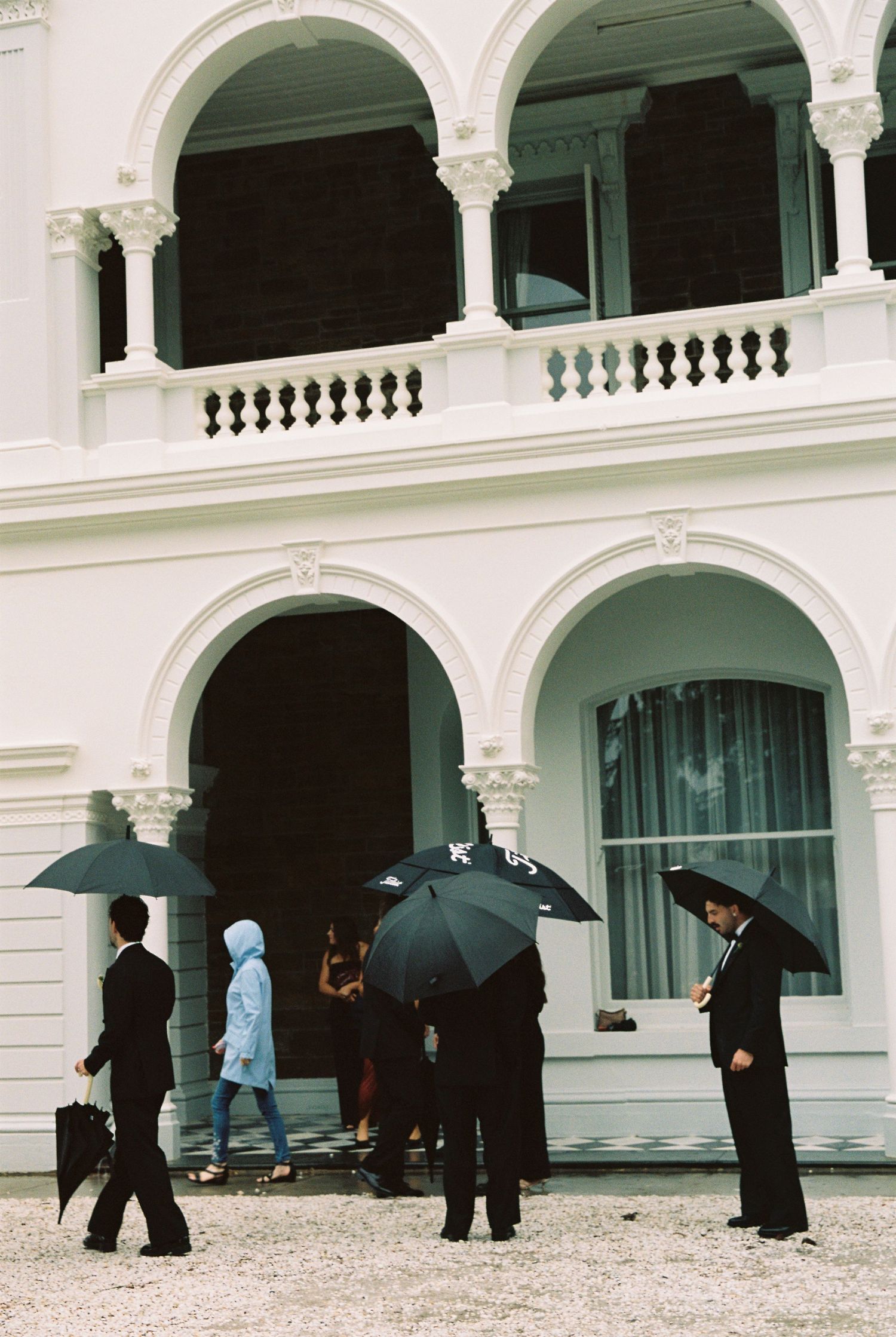 Wedding guests shelter under black umbrellas outside a grand white colonial-style building on a rainy day.