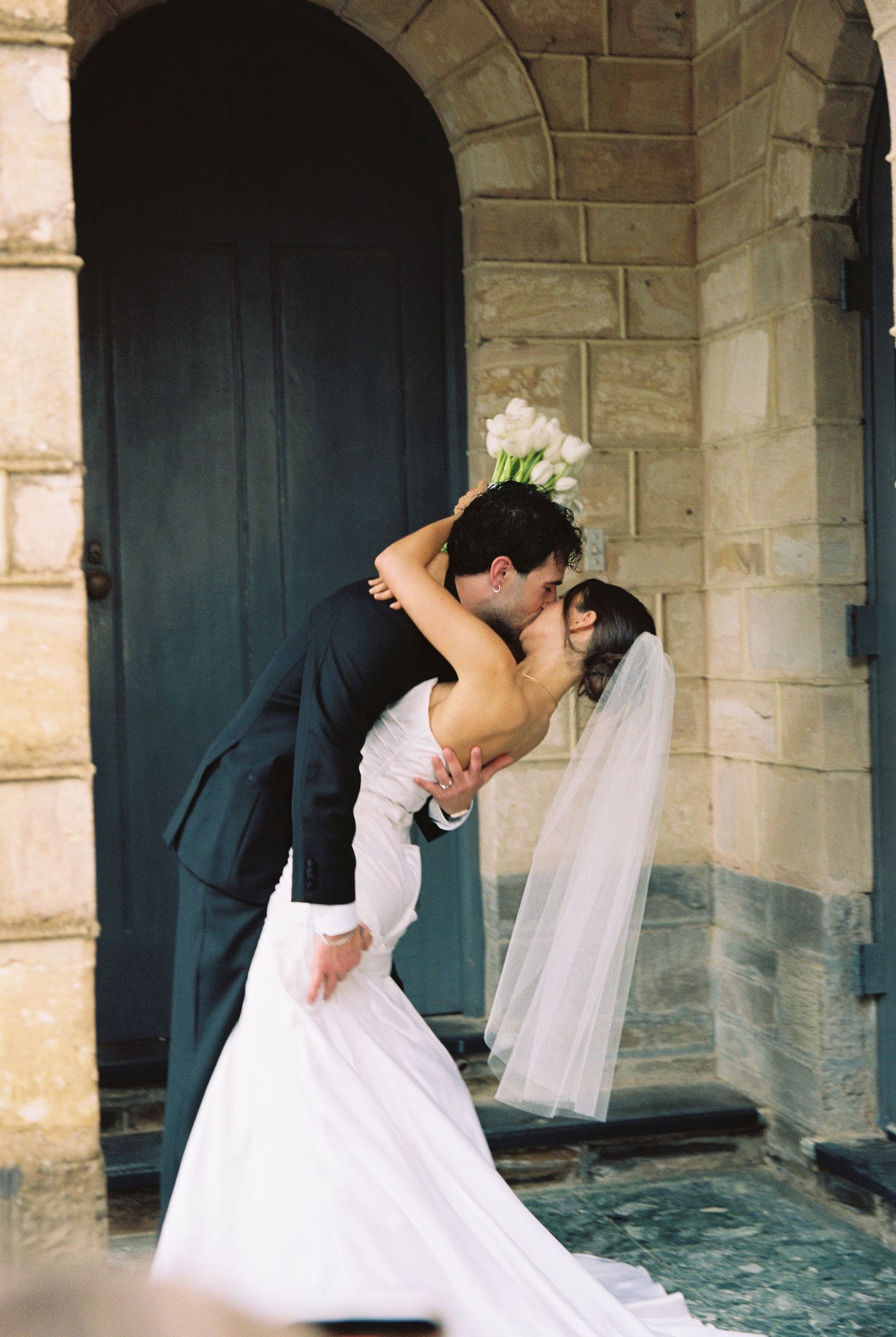 A bride and groom share a romantic dip kiss in front of a stone church doorway on their wedding day.