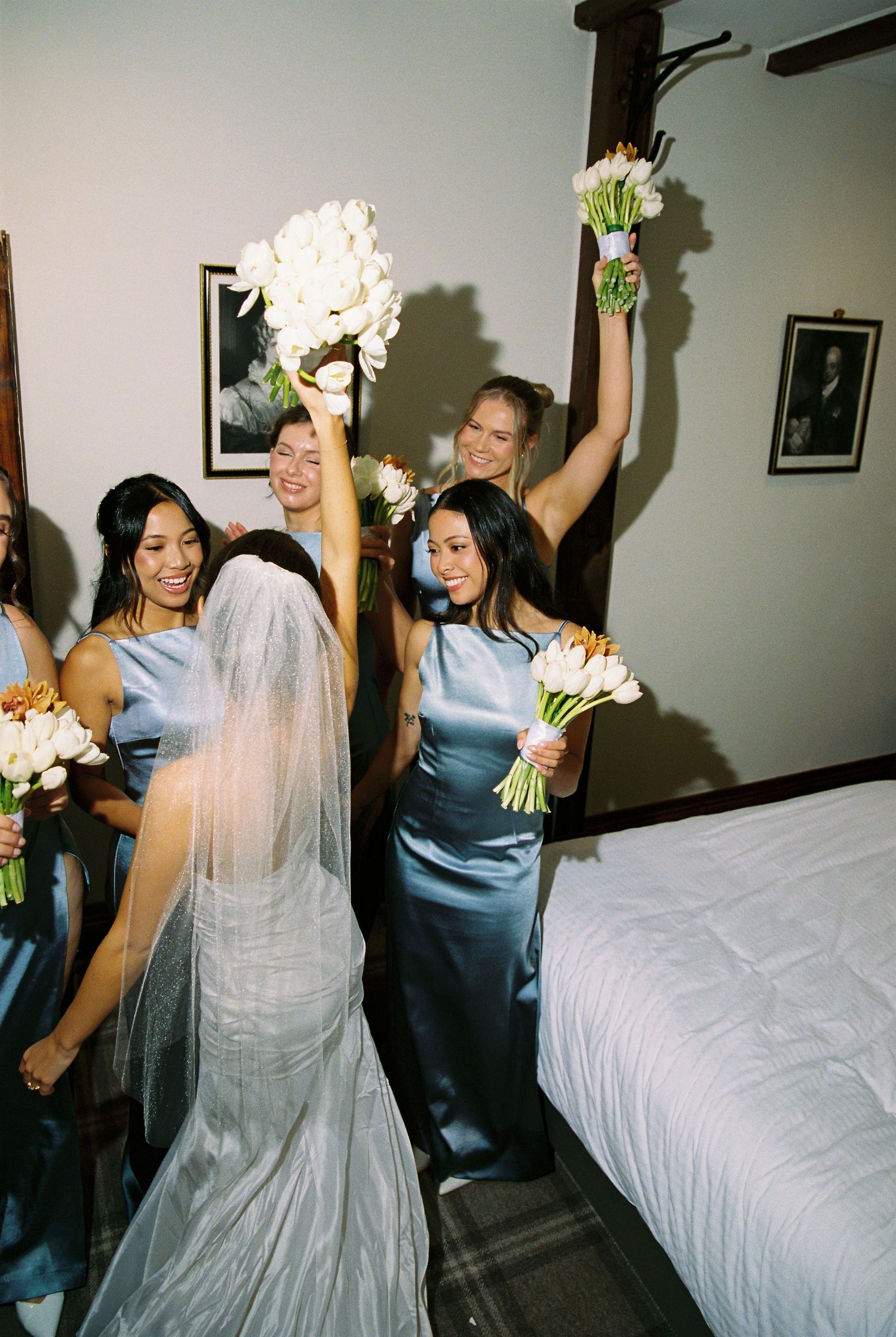 Excited bridesmaids in blue satin dresses raise white tulip bouquets celebrating with bride.