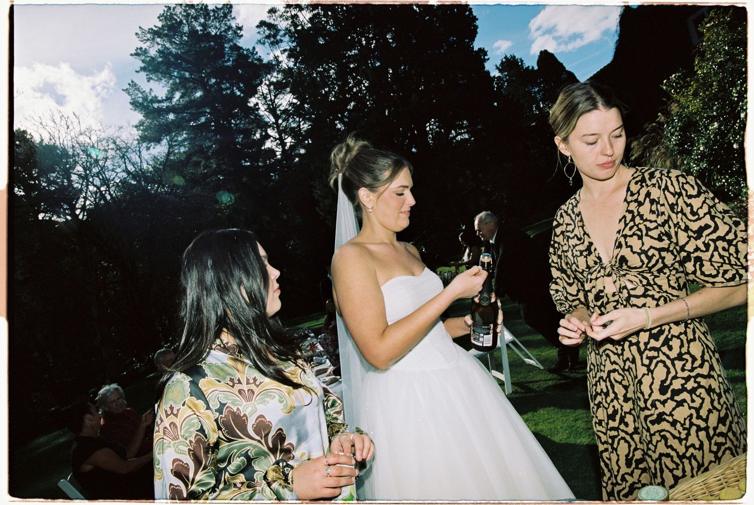 Bride in white gown celebrates with guests holding champagne bottles at outdoor wedding reception.