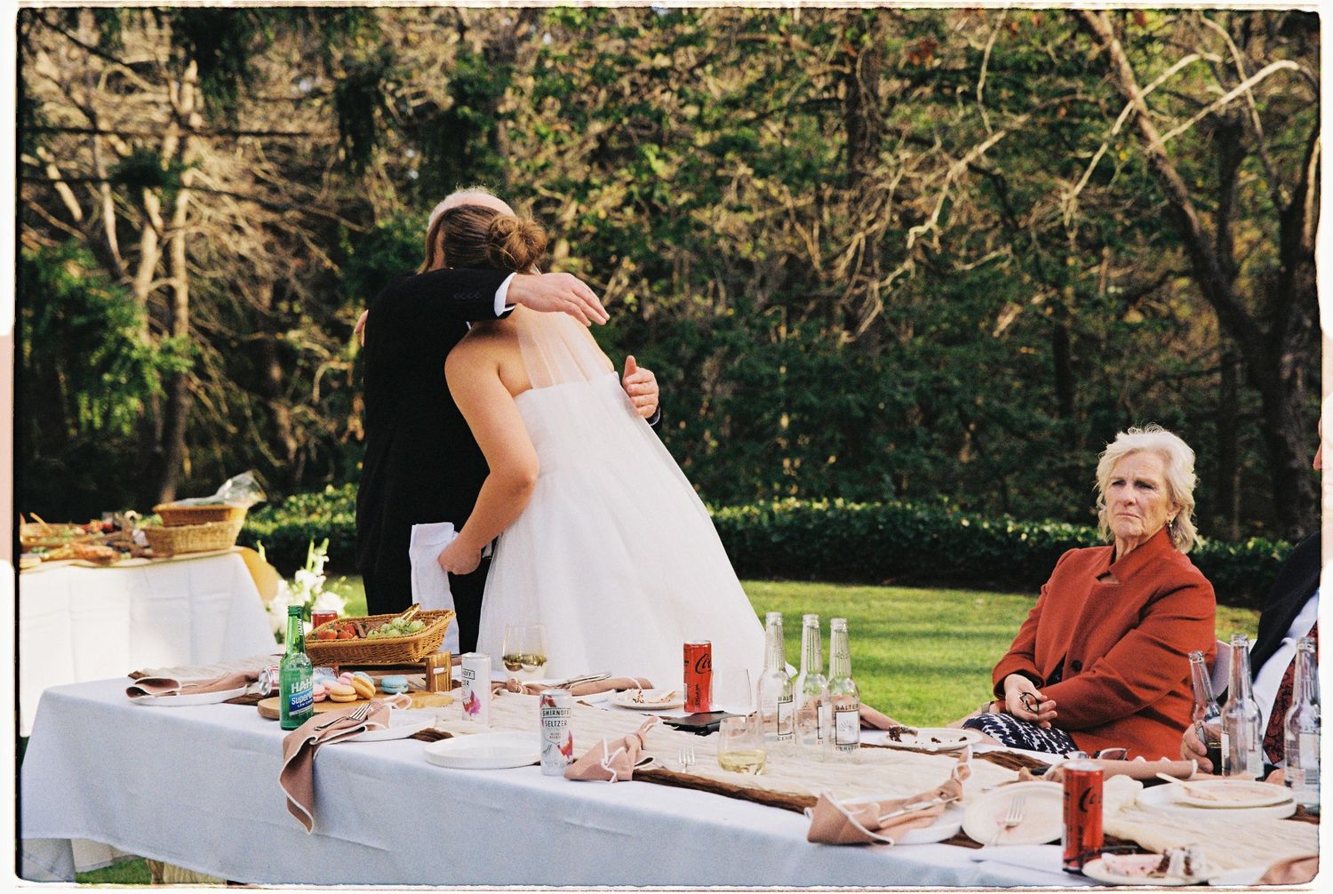 Bride and groom embracing at outdoor wedding reception table with elderly guest seated nearby in lush green garden setting.