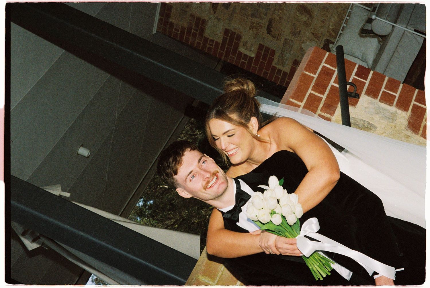 Bridesmaid in black dress holding white bouquet poses with groomsman on staircase.