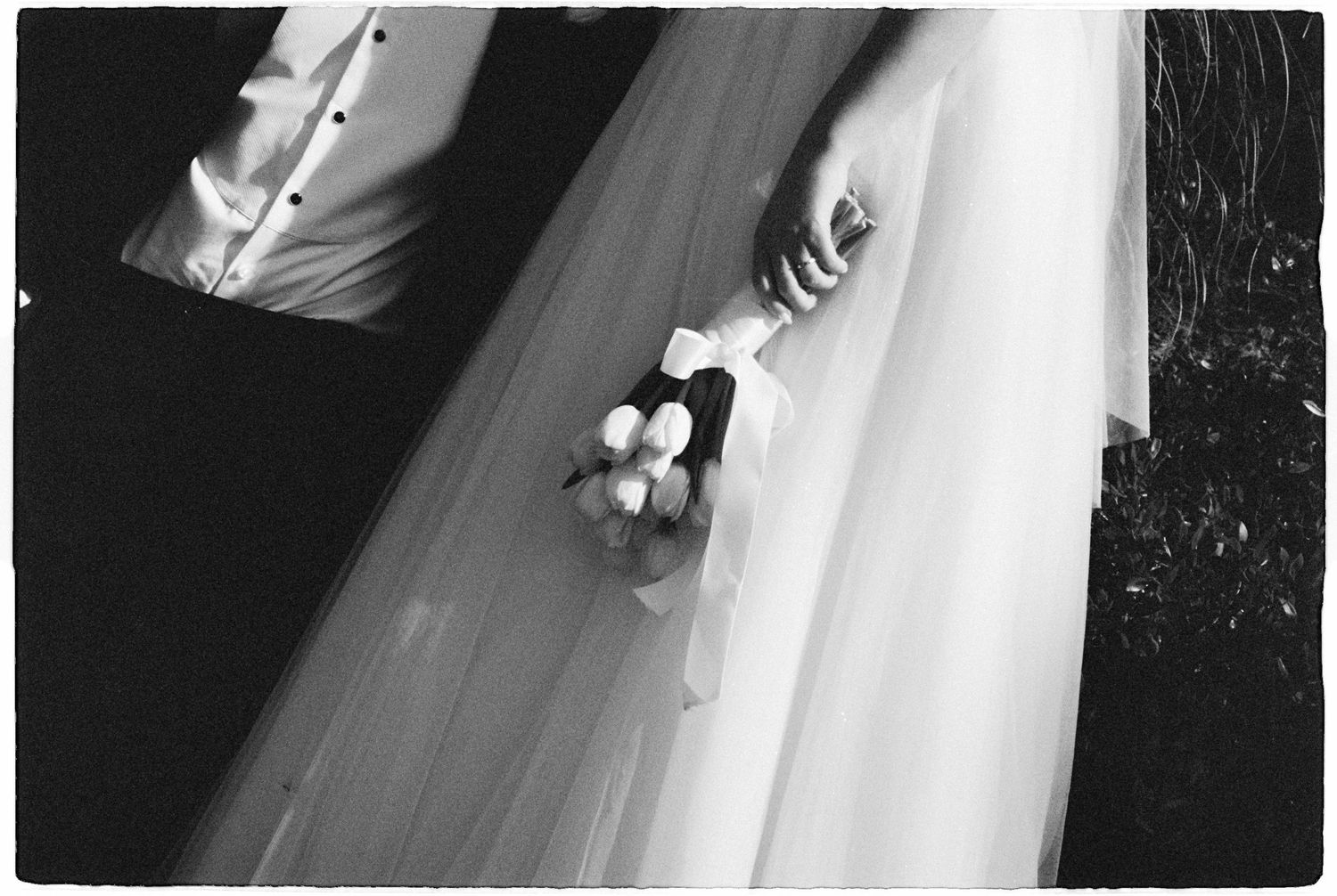 Black and white close-up of a bride holding a small floral bouquet with flowing veil cascading beside her groom.