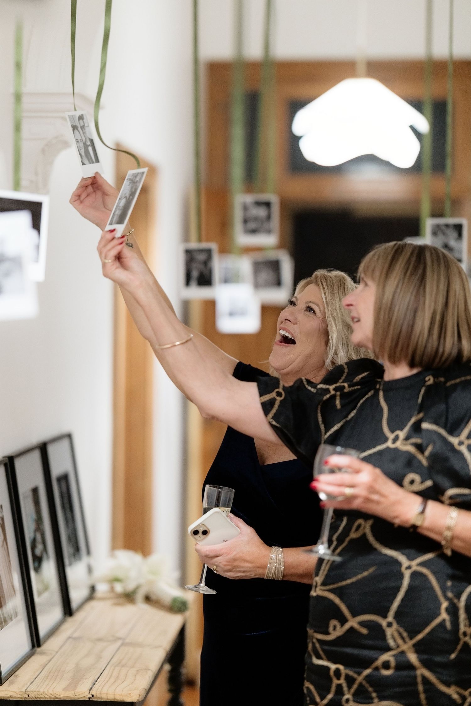 Two women in black dresses laugh while examining black and white photos hanging on a wall at a wedding reception.