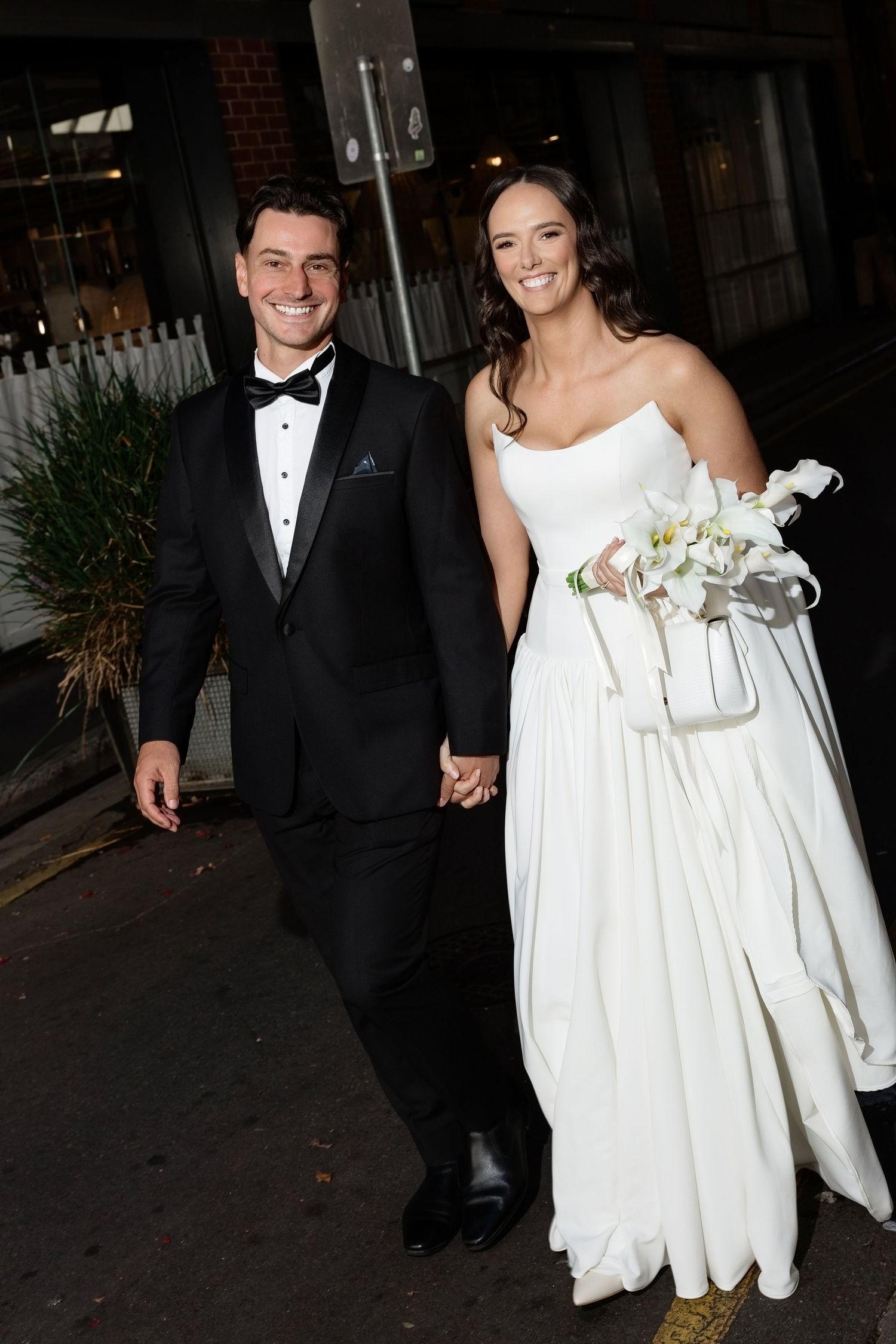 Happy bride and groom holding hands outdoors at night, bride carrying a white floral bouquet.