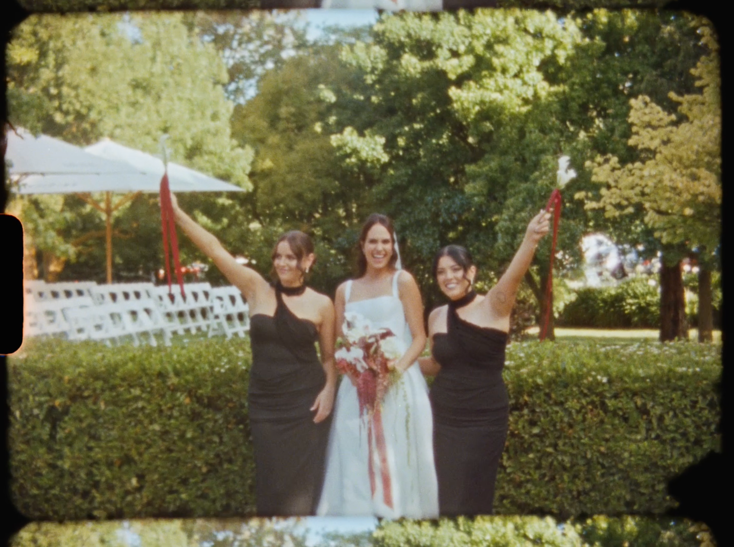 Bride with bouquet celebrates between two bridesmaids in black dresses raising ribbon wands in garden.