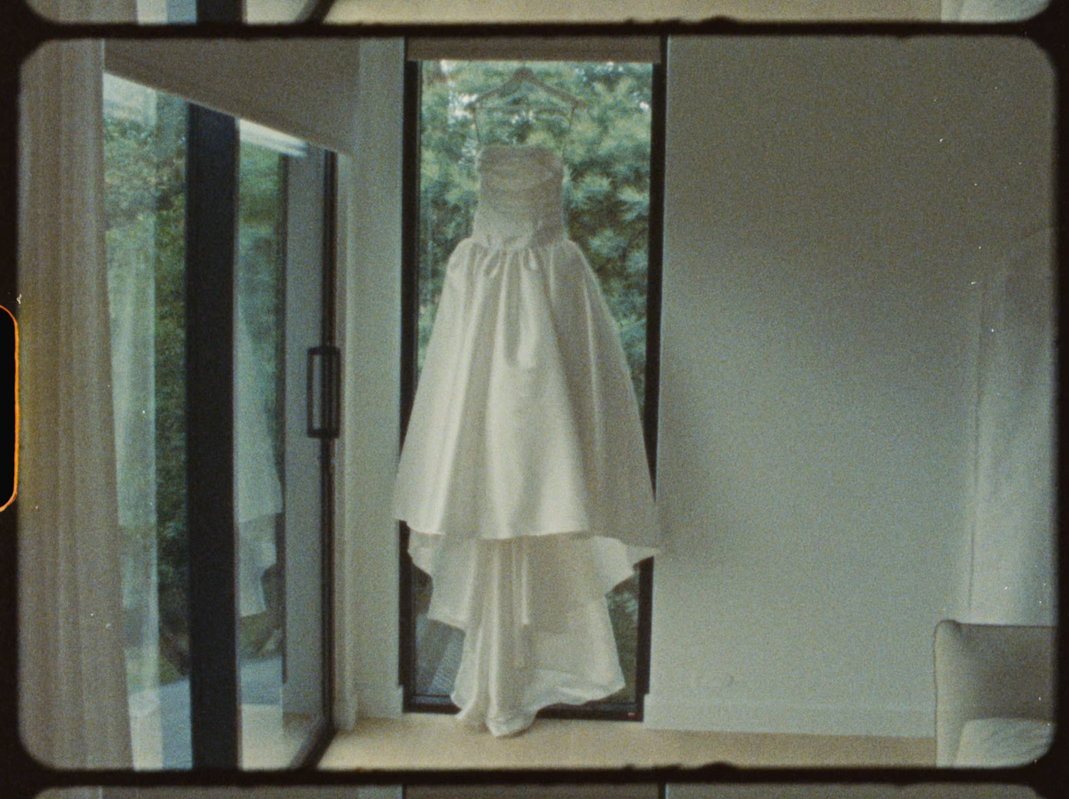 Full-length white wedding dress with layered skirt hanging in a sunlit doorway.