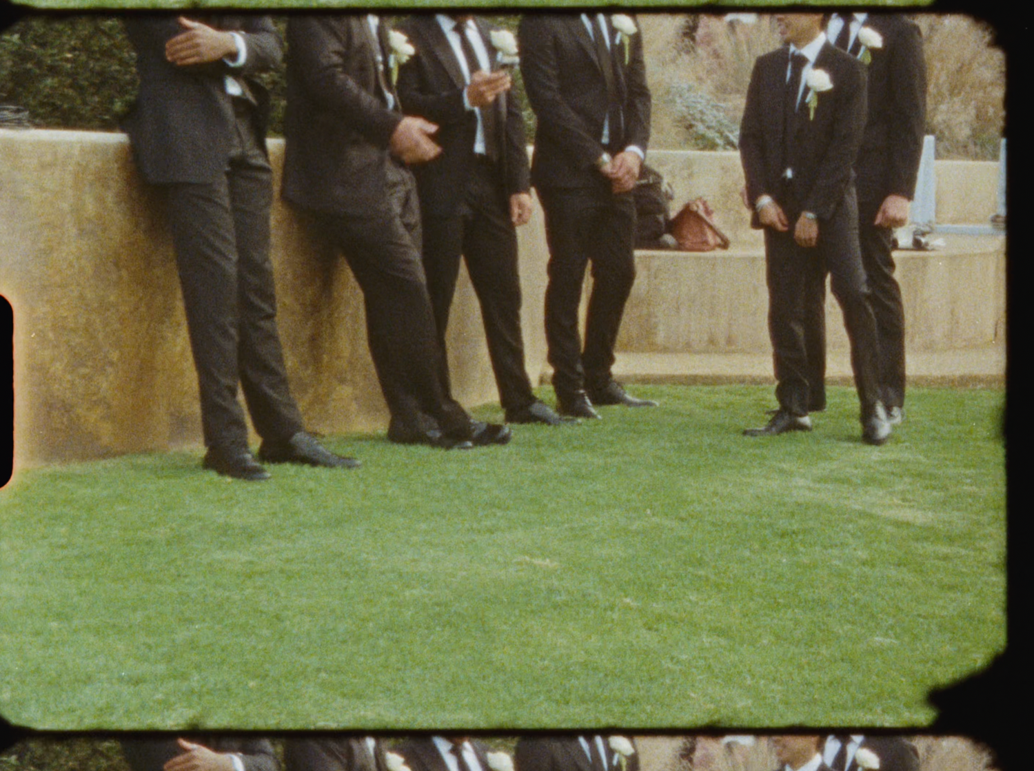 Groomsmen in matching black suits stand along stone wall on green lawn at outdoor wedding ceremony.