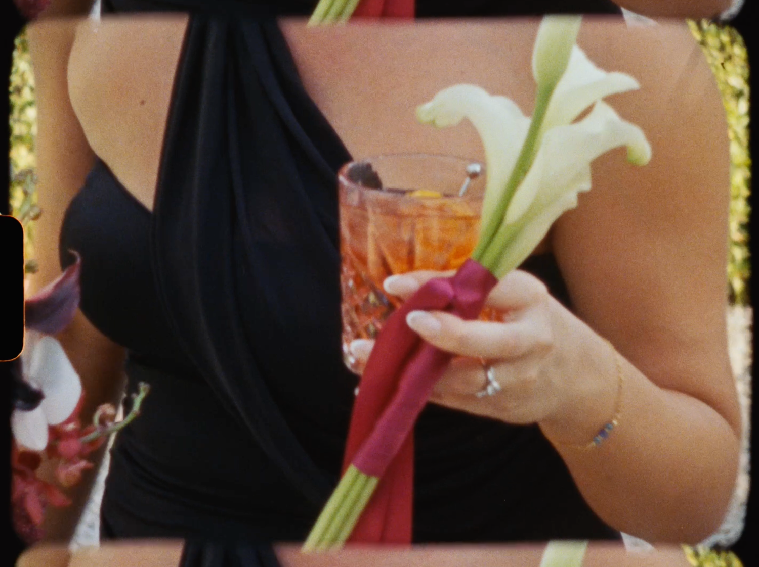 Bridesmaid in black dress holds white calla lily bouquet with red stems and an amber cocktail glass.