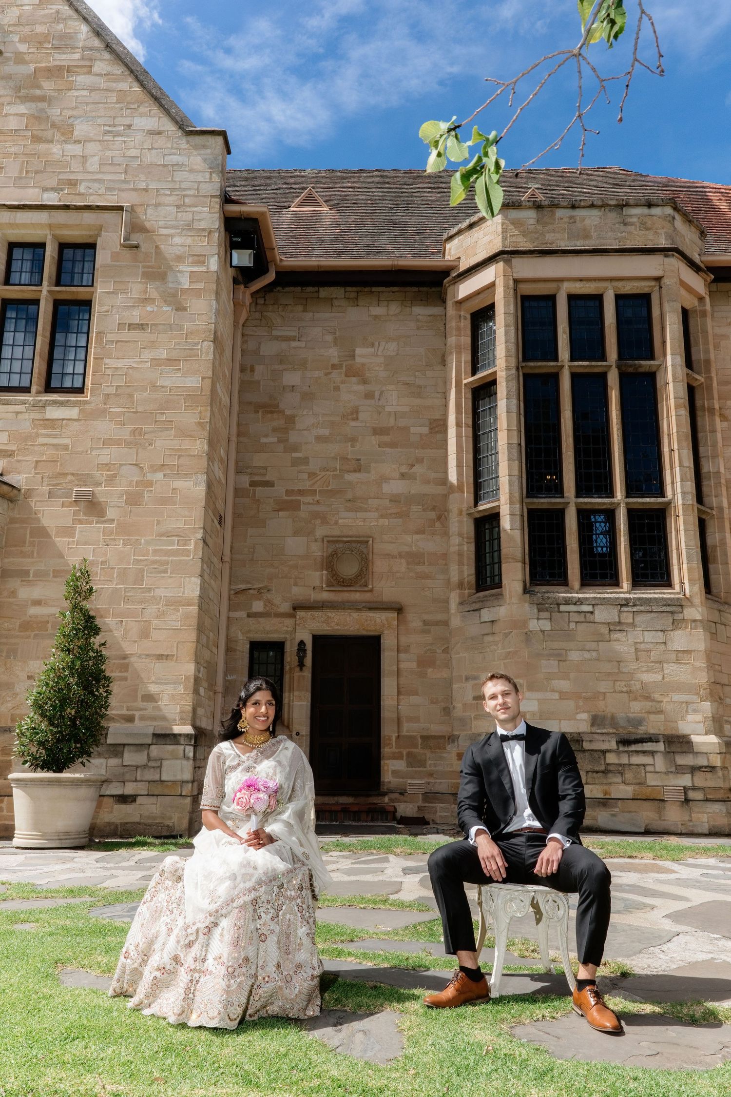 Bride and groom seated separately on chairs outside grand historic sandstone mansion on their sunny wedding day.