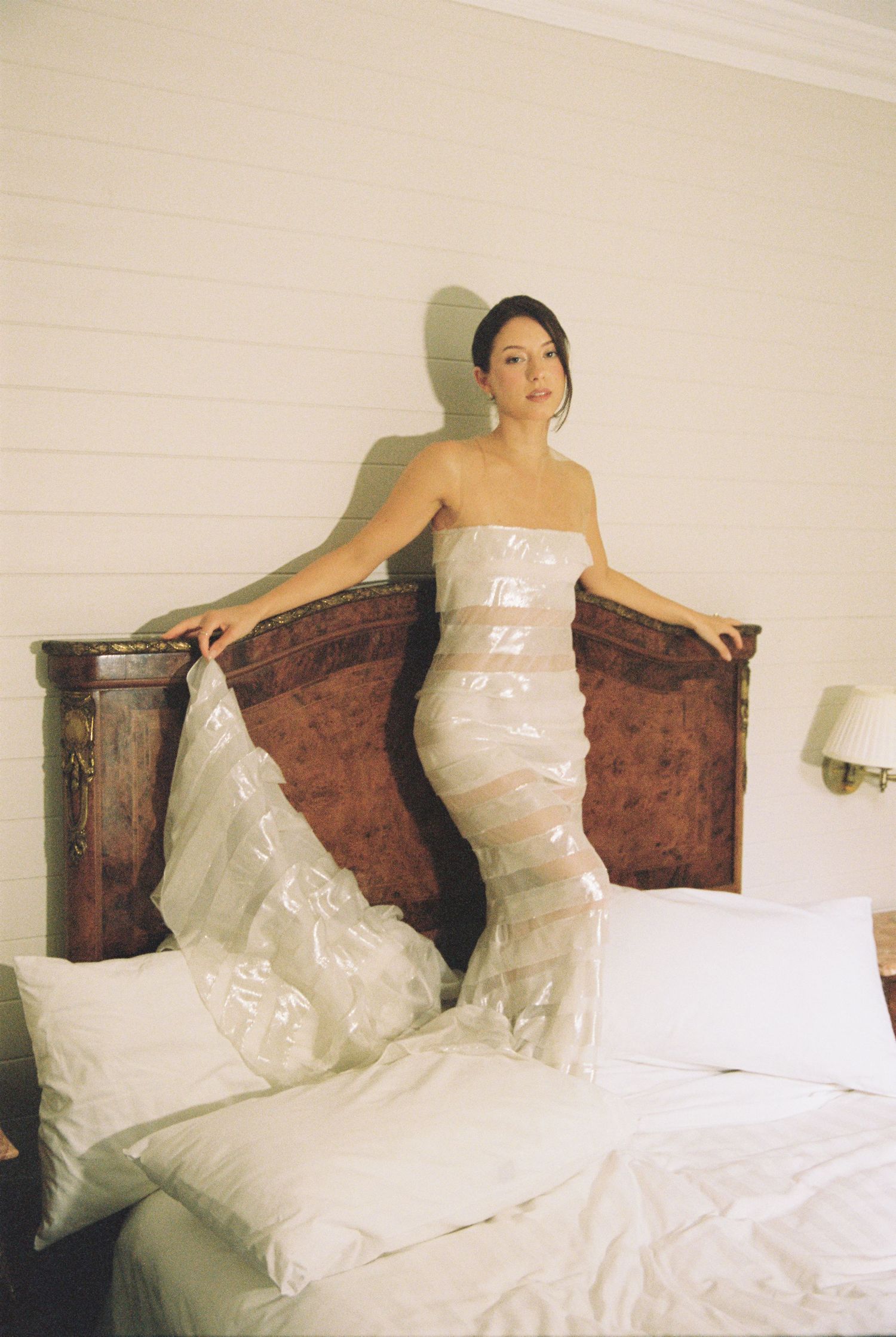 Bride standing on bed wrapped in wedding dress plastic garment bag before ceremony.