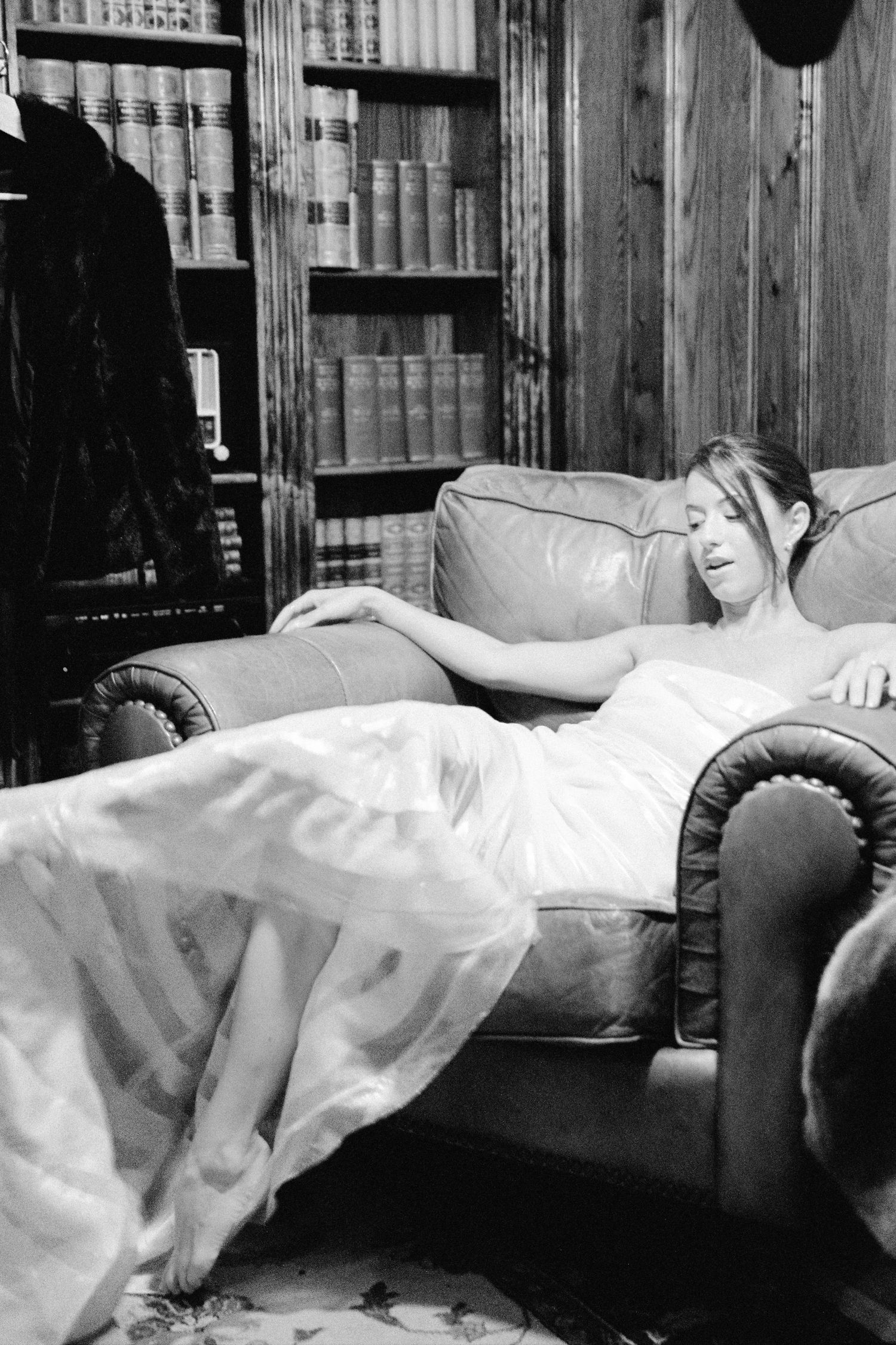 Bride in white gown reclining on leather sofa in wood-paneled library room.