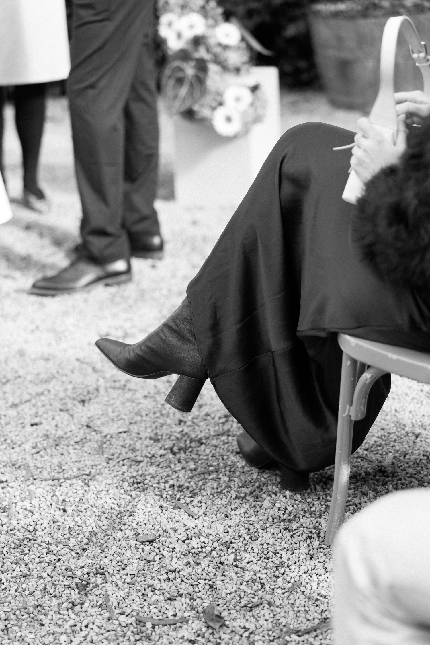 Black and white photo of a seated person in dark clothing and boots, smoking, with standing guests in background.