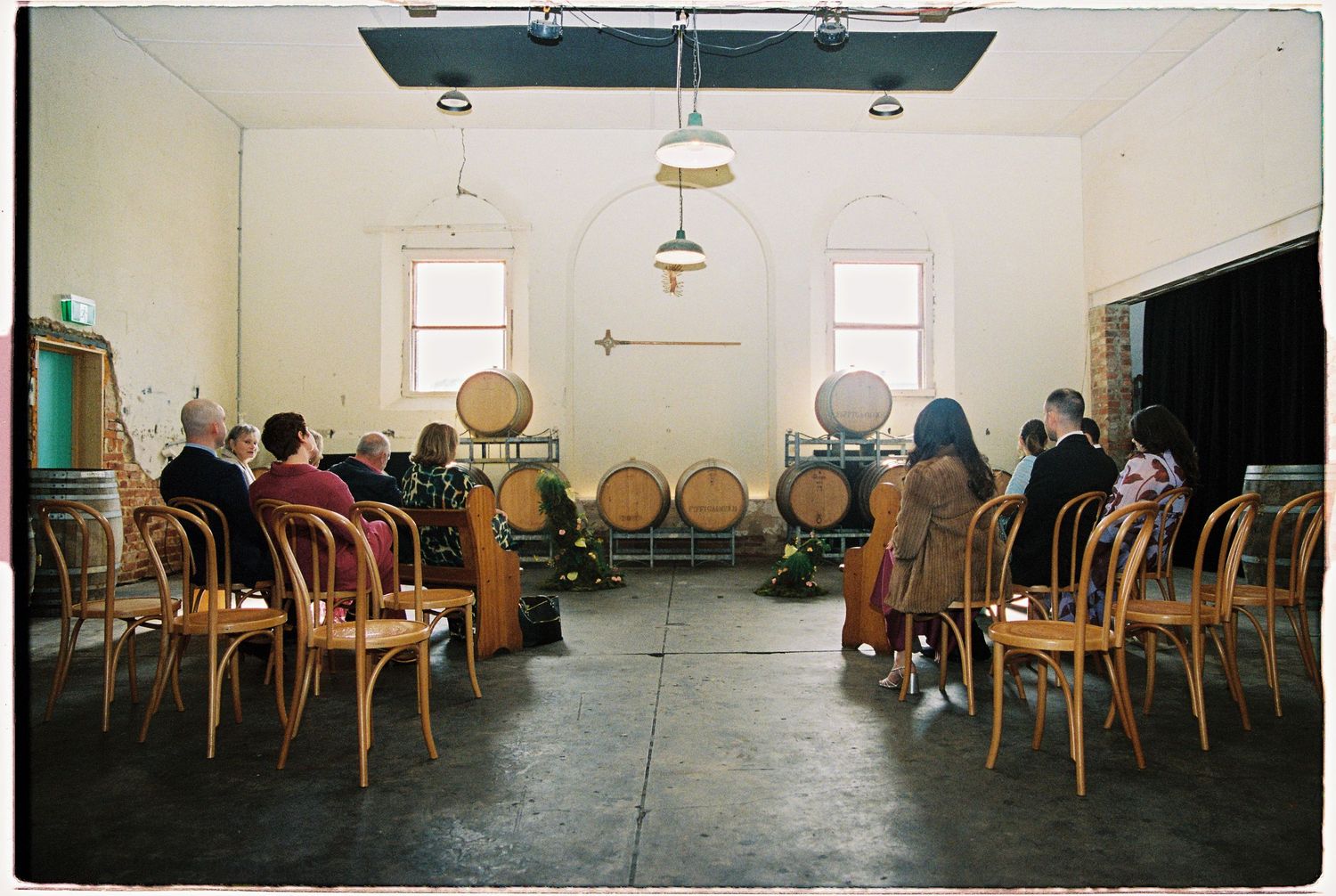 People seated in wooden chairs facing wine barrels in a rustic winery tasting room with pendant lights.